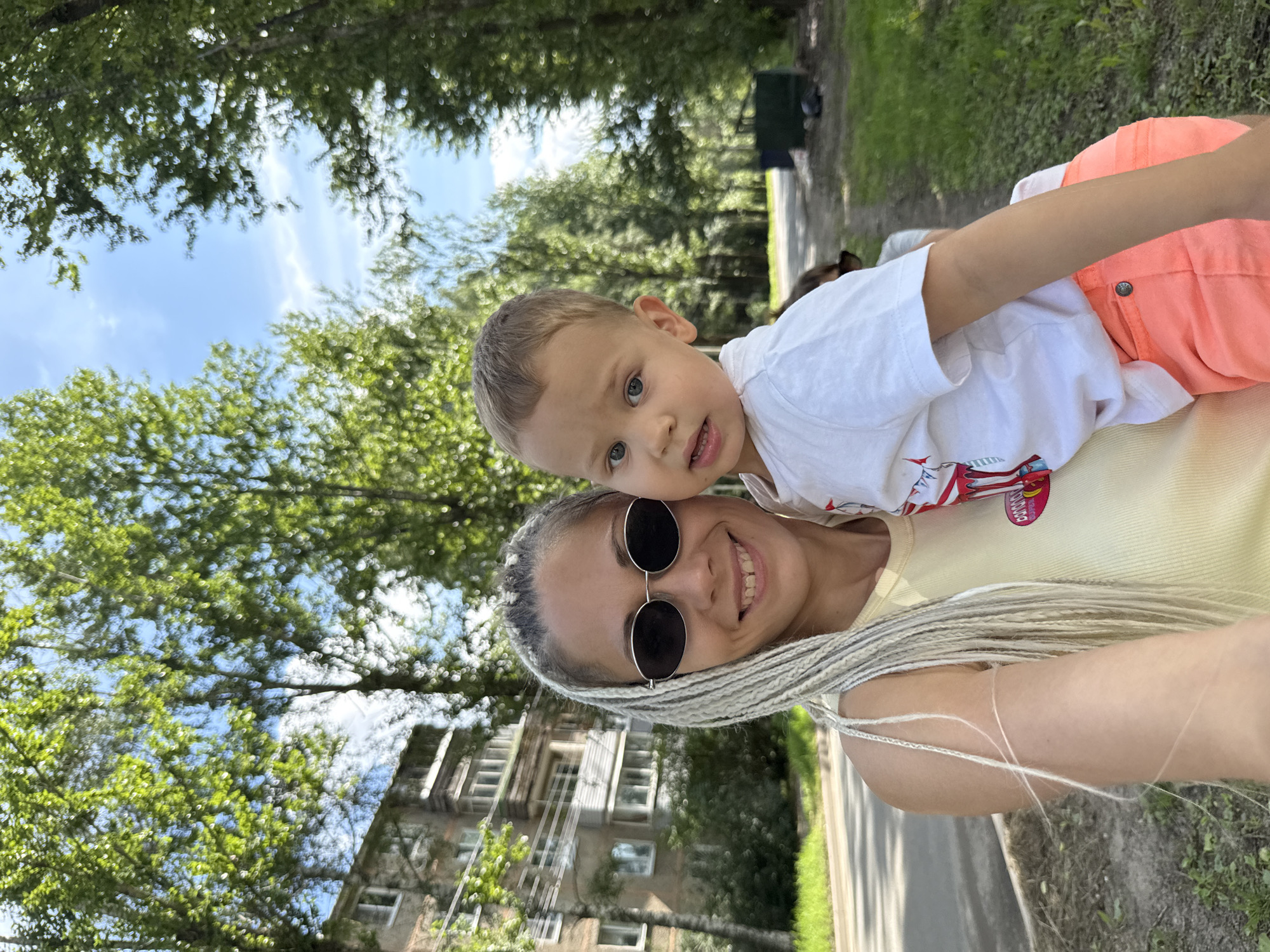 In Rusanovo, Russia, a woman with long, blonde braids and sunglasses holds a young boy. The woman wears a yellow tank top and is smiling at the camera. The boy, wearing a white t-shirt with a colorful graphic and coral-colored shorts, looks towards the camera with a curious expression. They are positioned in front of a tree-lined street, with a brick apartment building visible on the left. The background shows more trees and a blue sky with scattered clouds, suggesting an outdoor setting.