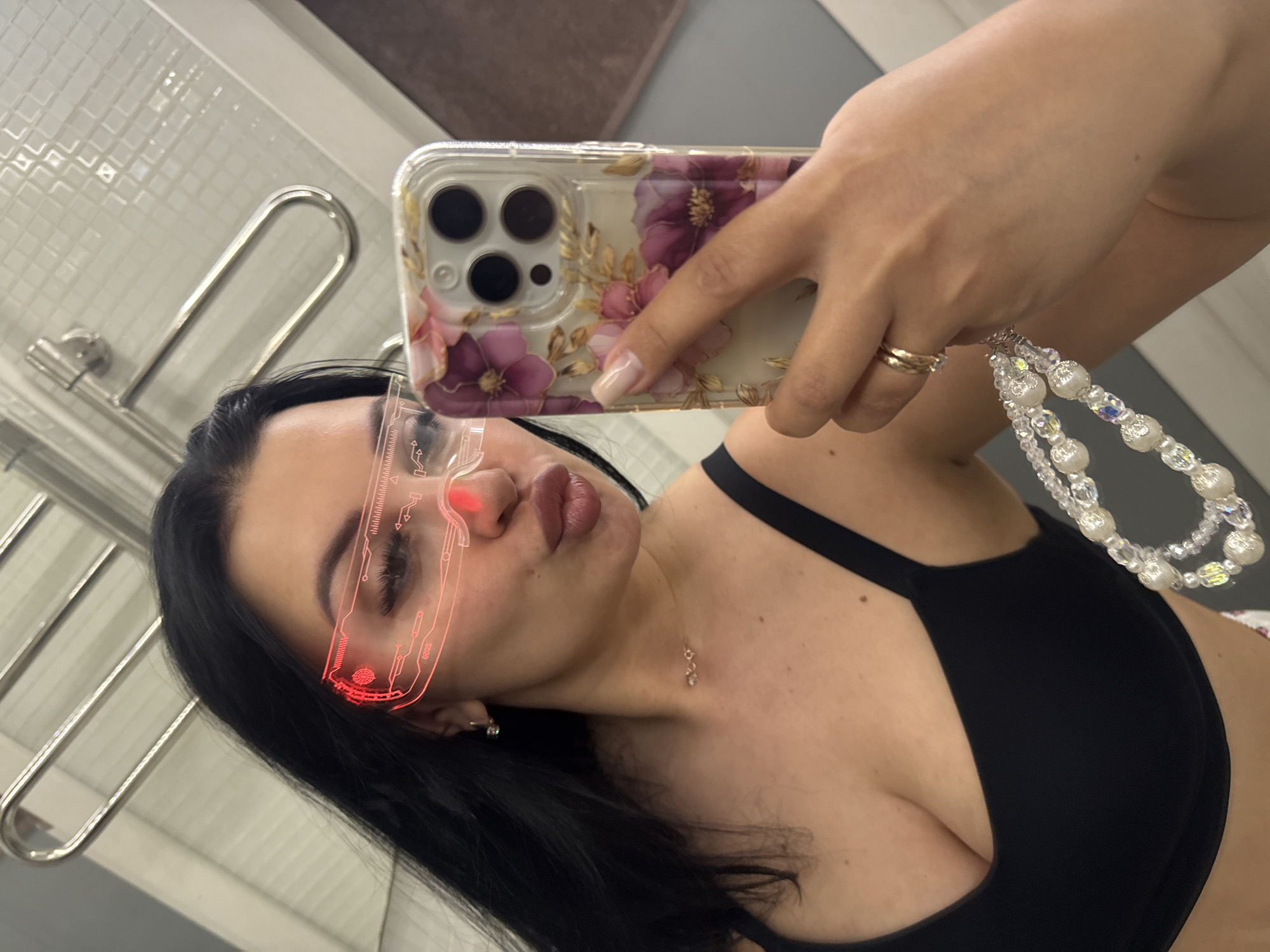 A woman is taking a selfie in what appears to be a bathroom. She is wearing a black top and has a clear phone case decorated with flowers. The phone has a beaded wrist strap. She is also wearing a gold ring. A digitally added red neon-like graphic is superimposed on her face, resembling futuristic glasses. The background includes a white tiled wall and a chrome towel warmer. There is no information to suggest this image was taken in Vladivostok, Russia.