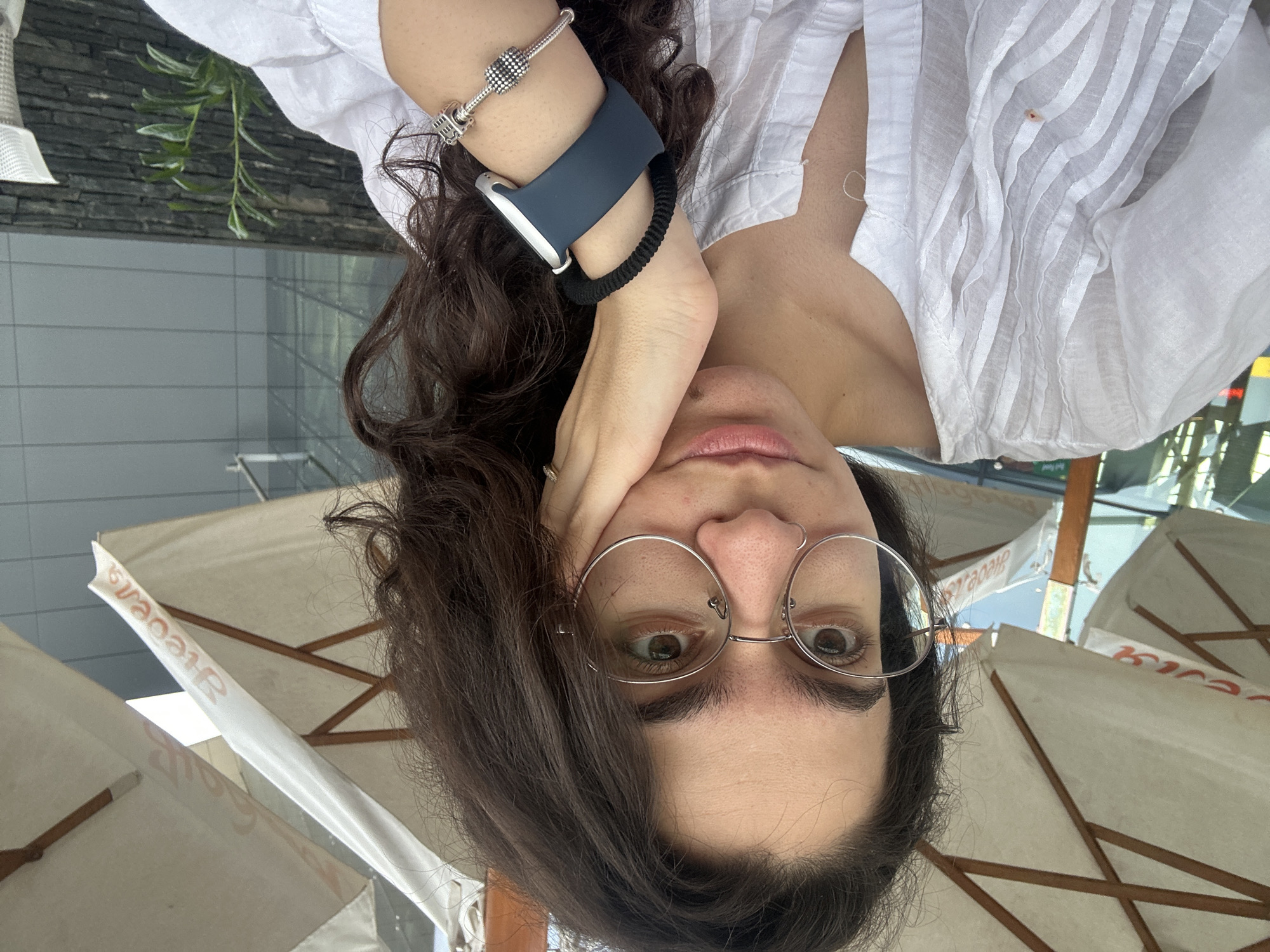 A young woman with dark, curly hair and glasses is captured in a close-up, frontal selfie. She is wearing a white, textured blouse and resting her left hand on her cheek. A smart watch with a dark blue band is visible on her left wrist, along with a silver charm bracelet. She is outdoors, likely at a cafe, with a white umbrella and wooden supports of an outdoor canopy visible in the background. The environment suggests a sunny day in an urban setting. The image is composed in a way that focuses on the woman's face and upper body, with the background slightly blurred.