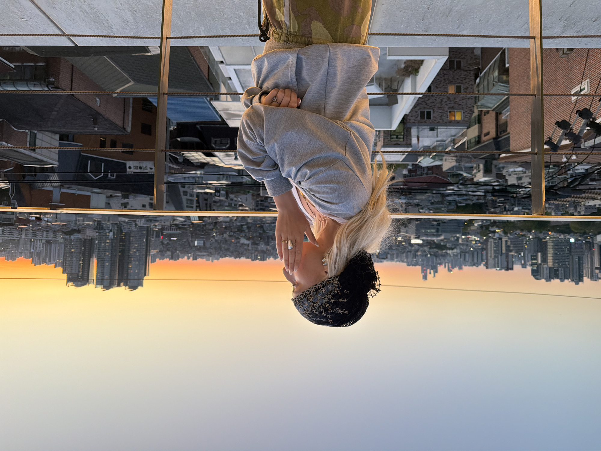 The image depicts a woman in front of a cityscape at dusk. The woman is wearing a grey hoodie, camo pants, and a black head covering with floral detailing. Her blonde hair is visible. She is holding her hand up to her face. She is standing behind a railing. In the background is a sprawling urban landscape with numerous buildings silhouetted against the sunset sky. The city is likely Seoul, South Korea. The buildings closer to the railing appear to be residential buildings, while those farther away appear to be skyscrapers. The sky transitions from a pale blue at the bottom of the frame to a vibrant orange near the horizon. The image is upside down.