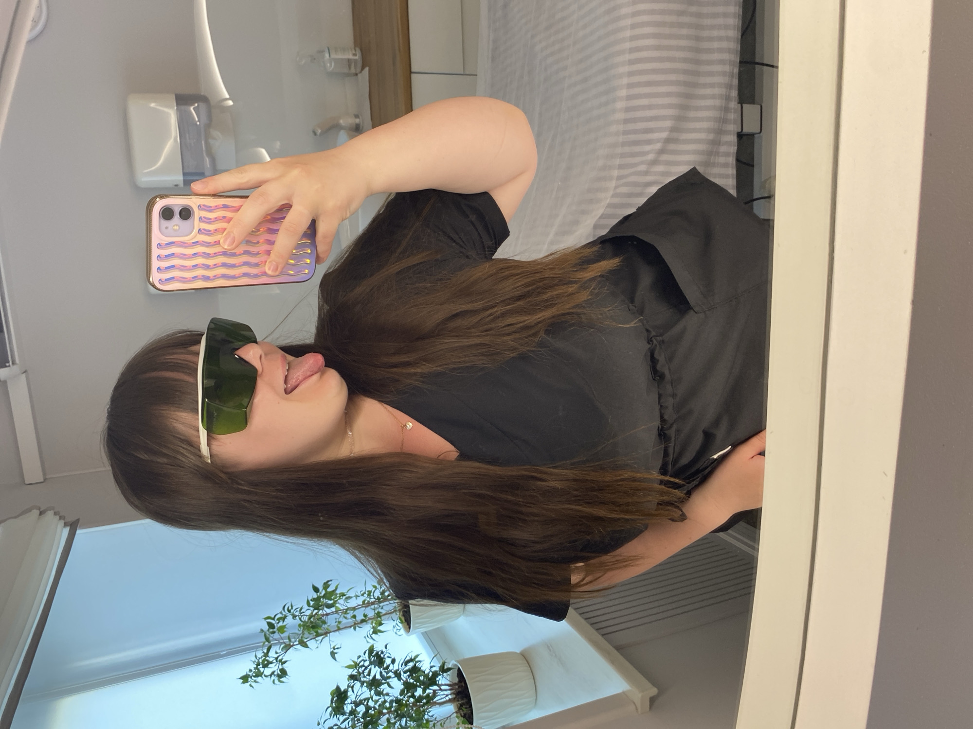 A woman is taking a selfie in a mirror. She is wearing protective eyewear and sticking her tongue out. She is holding a cell phone with a patterned case. The background includes what appears to be a dental or medical office, suggested by the equipment visible, and a room with a striped bedspread. There are potted plants visible near a window. The location cannot be verified as Votkinsk, Russia from the image.