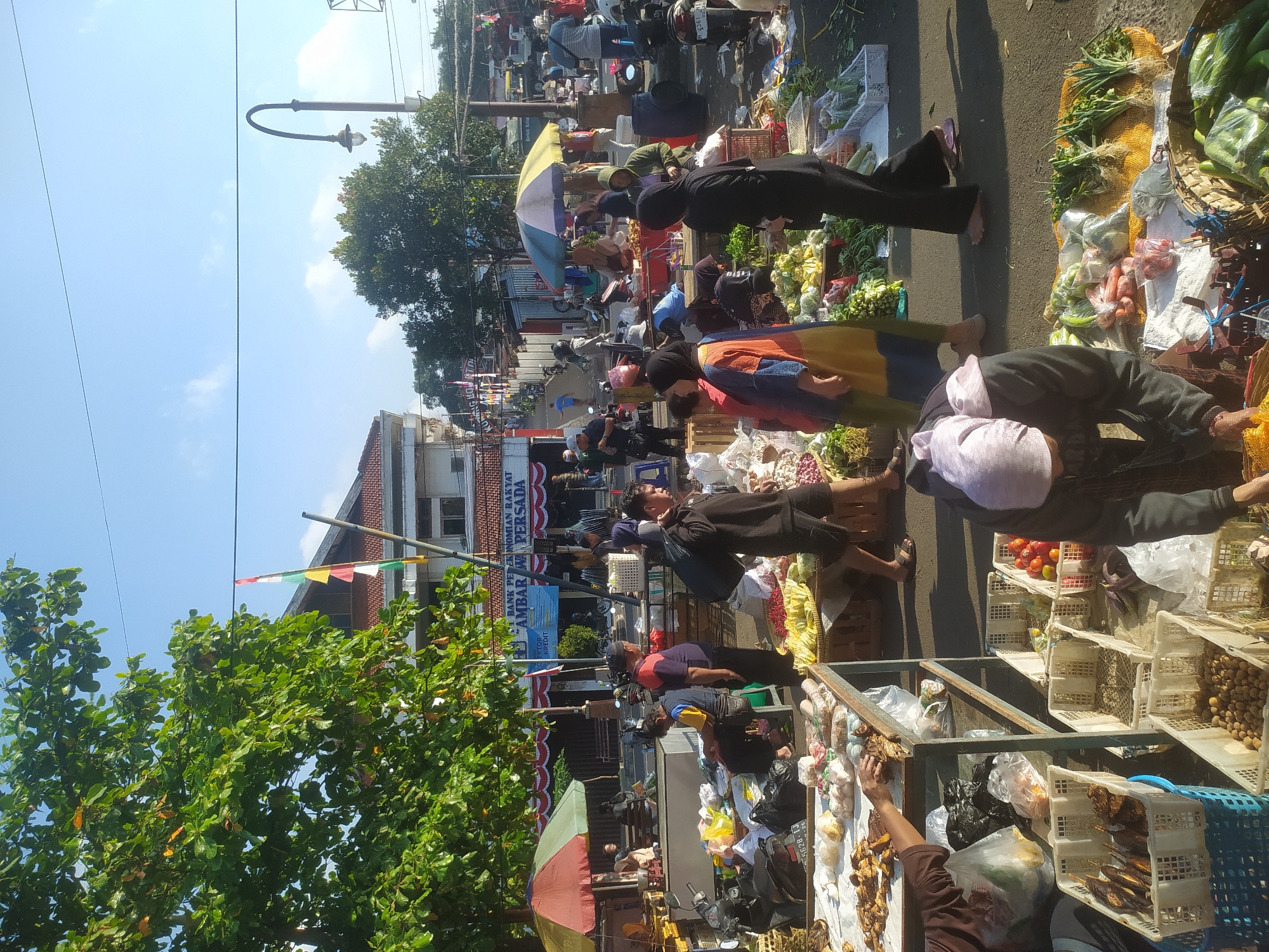 This image depicts a bustling outdoor market scene in Kupang, Indonesia, likely in a public square or along a street with market stalls. The time appears to be morning, given the clear blue sky and the quality of the sunlight casting defined shadows.
In the foreground, several market stalls are visible, laden with a variety of fresh produce. Baskets overflow with vegetables such as green beans, cucumbers, carrots, and leafy greens, alongside red tomatoes and what appear to be onions or shallots. One stall features dried fish or meat. Various plastic bags, crates, and woven baskets are used to display and transport the goods.
A number of individuals are engaged in activities related to the market. Several people are dressed in traditional Indonesian attire, including hijabs and long-sleeved shirts. Some are vendors attending to their stalls, while others appear to be shoppers browsing the merchandise. One person in the immediate foreground, wearing a dark hooded jacket and a light-colored headscarf, is bending over a stall, possibly attending to the produce. Another person to their right, in black pants and a black headscarf, is walking away from the camera. In the mid-ground, a man in a blue vest and a man in a purple shirt are standing near a seated individual. People are also visible in the background, interacting at other stalls and moving through the market.
The environment includes a street or paved area where the market is set up. In the background, there are buildings, including one with a red tile roof and a sign that reads "BANK PERKONOMIAN RAKYAT AMBAR WA PERSADA," indicating a financial institution. A lamppost with an ornate fixture stands near the right side of the image. Trees with dense green foliage are scattered throughout the scene, providing some shade. Flags are visible on poles in front of one of the buildings. Motorcycles are parked in various locations, suggesting this is a common mode of transportation. The overall impression is one of a lively and active local marketplace.