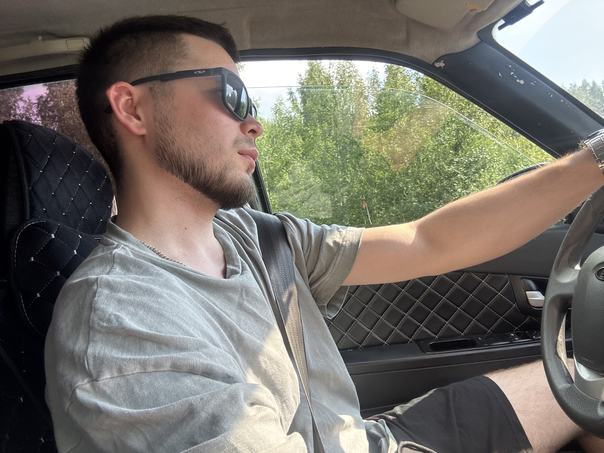 In the city of Marchugi, Russia, a young man wearing sunglasses and a grey t-shirt is driving a car. He has a beard and short hair. The car's interior shows a black upholstered seat and a leather-wrapped steering wheel. Outside the car, green trees are visible through the window. The sunlight casts shadows on the man's face and shirt.