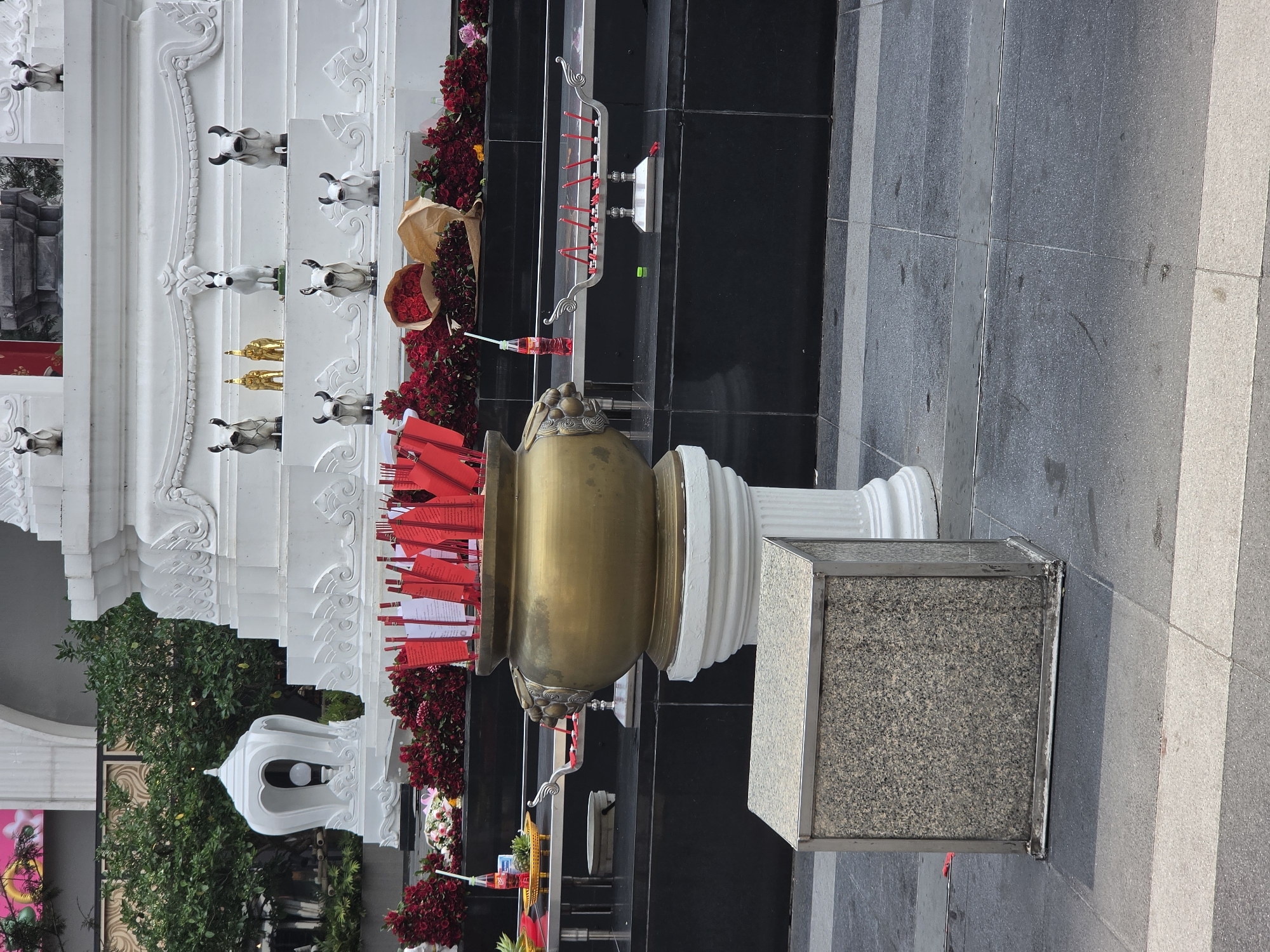 The image depicts a shrine in what is likely Bangkok, Thailand, based on architectural and cultural elements. A large brass incense pot overflowing with red and white sticks dominates the foreground. The pot sits atop a white column and a grey stone base.
Behind the pot is a multi-tiered white structure with decorative carvings and animal statues. Red roses and other offerings are arranged on the structure. A black wall forms a backdrop.
The setting appears to be an urban outdoor space, with grey pavement visible in the foreground and on the right. In the periphery, there are glimpses of greenery and a building, suggesting a busy area within the city.