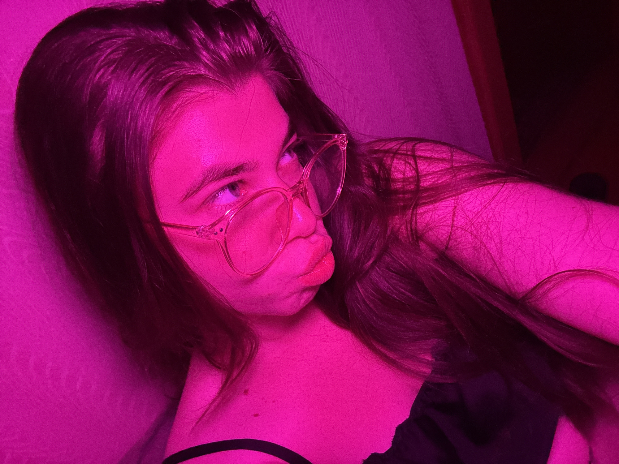 A young woman with long, dark hair is captured from a close-up perspective, her face angled away from the camera. She is wearing glasses with clear, thin frames. The lighting is predominantly pink, casting a vibrant hue over the entire scene. Her hair falls loosely around her shoulders and across her upper body, partially obscuring her clothing, which appears to be a dark-colored top with thin straps. The background is a textured surface, likely a wall, illuminated by the same pink light. The image offers a glimpse of her profile, with her eyes and lips slightly visible.