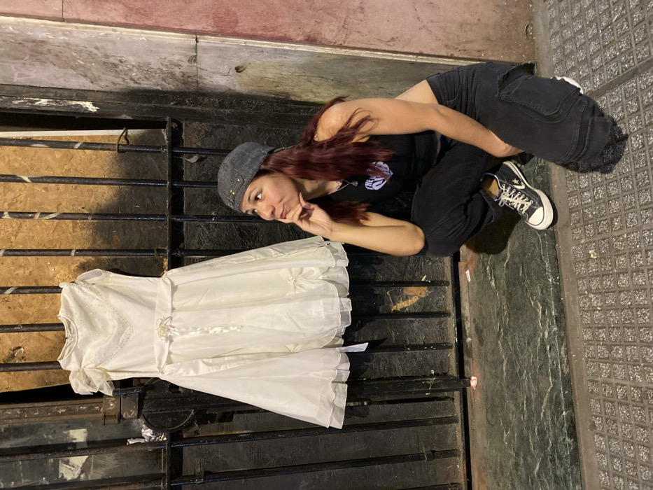 A crouching young woman in a gray cap, black top, and black pants looks pensively at a white dress hanging on a black barred gate. She rests her chin on her hand in contemplation. The gate appears to be part of a building's facade. She wears black sneakers, and the ground around her is tiled. The scene seems to be outdoors, possibly at night, given the lighting.