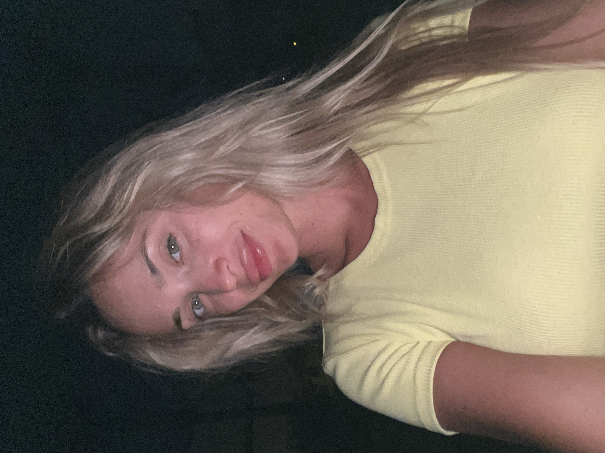 This is a close-up, head-and-shoulders selfie of a young woman with blonde hair and light-colored eyes. She is wearing a light yellow, ribbed t-shirt. Her head is tilted slightly to her left, and she is looking directly at the camera with a slight smile. The background is dark, suggesting it is nighttime. The image appears to have been taken outdoors, possibly in a garden or on a patio, as indicated by a faint out-of-focus light source in the background on the right side. There is no visible indication of the specific location within Sharm El Sheikh, Egypt, or any other forensic details about the scene.