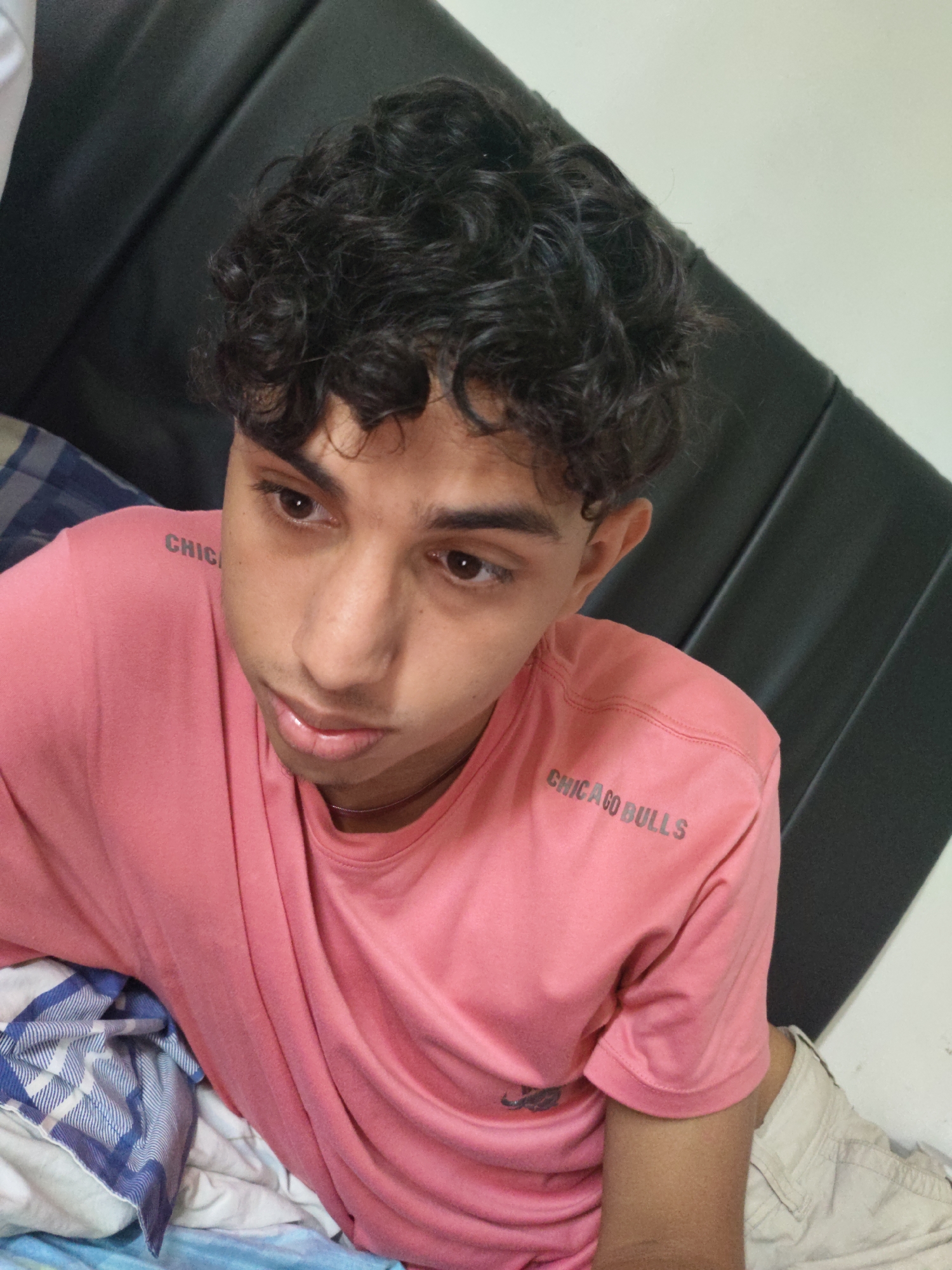 The image shows a close-up, high-angle shot of a young male, possibly a teenager, with curly dark hair. He is wearing a pink t-shirt with "CHICAGO BULLS" printed on the sleeve and beige shorts. The subject is looking down and to the right of the frame. He appears to be lying down, with a dark padded surface behind him, possibly a headboard or couch. A blue and white striped blanket is visible in the lower left corner of the image. The setting does not provide any visual cues to indicate a specific location in Piloncito, Venezuela.