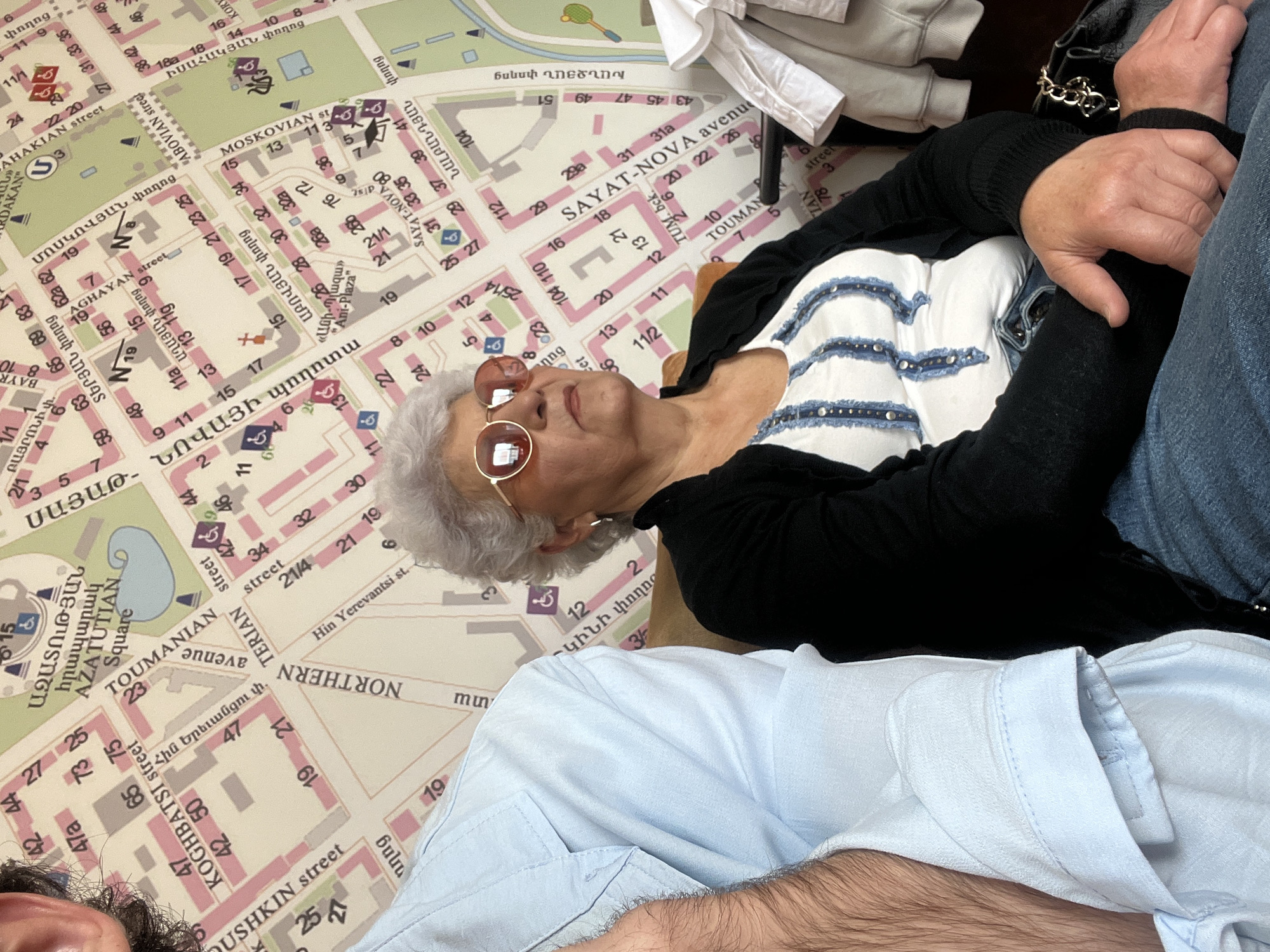 A high-angle shot features a woman in sunglasses and casual attire, partially obscured by two other individuals. The woman lies on a map, specifically a detailed rendering of Yerevan, Armenia. Text on the map indicates streets like Northern Avenue, Toumanian Avenue, Abovian Street, and Sayat-Nova Avenue, along with prominent locations like Azatutian Square. The immediate environment suggests an indoor setting, possibly a restaurant or cafe, given the casual positioning of the subjects and their placement on the map.