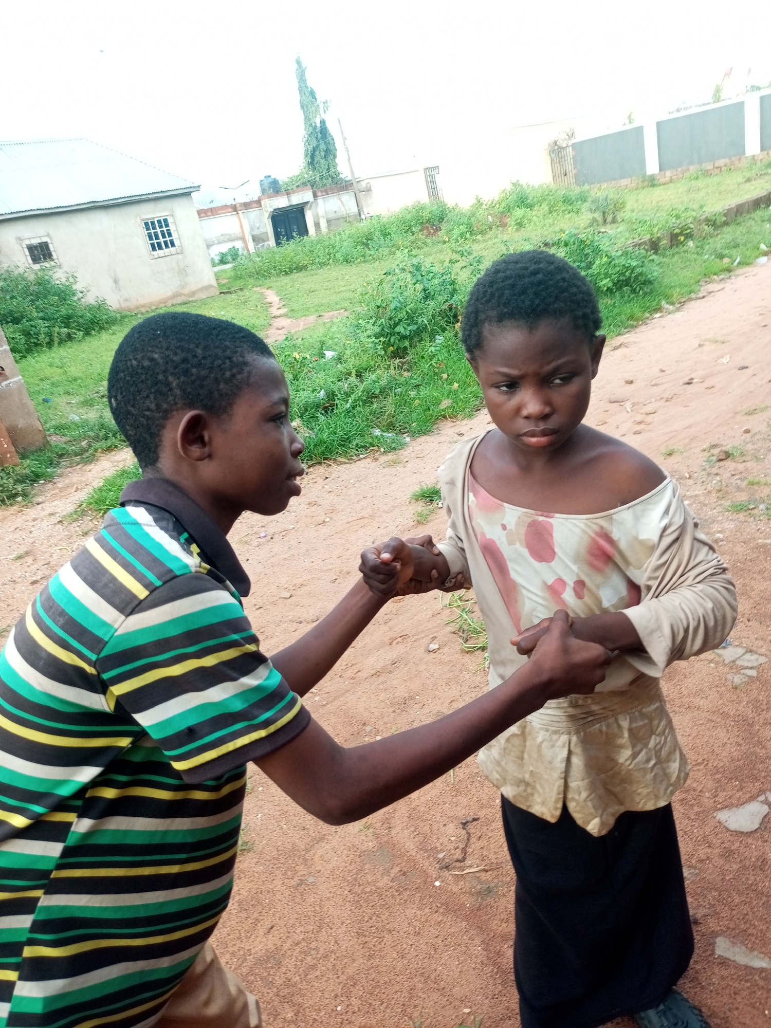 In Tempure, Nigeria, two children are captured in a close-up outdoor shot. A boy on the left, wearing a horizontally striped shirt in green, yellow, white, and black, faces a girl on the right. The girl is dressed in a cream-colored, long-sleeved shirt with reddish-pink splotches and a black skirt. Their hands are clasped together in the center of the frame, suggesting an interaction between them. The background reveals a dusty, reddish-brown path and sparse green vegetation. In the distance, a light-colored building with a metal roof and windows is visible, along with a fence and more greenery. The overall lighting suggests an overcast day.