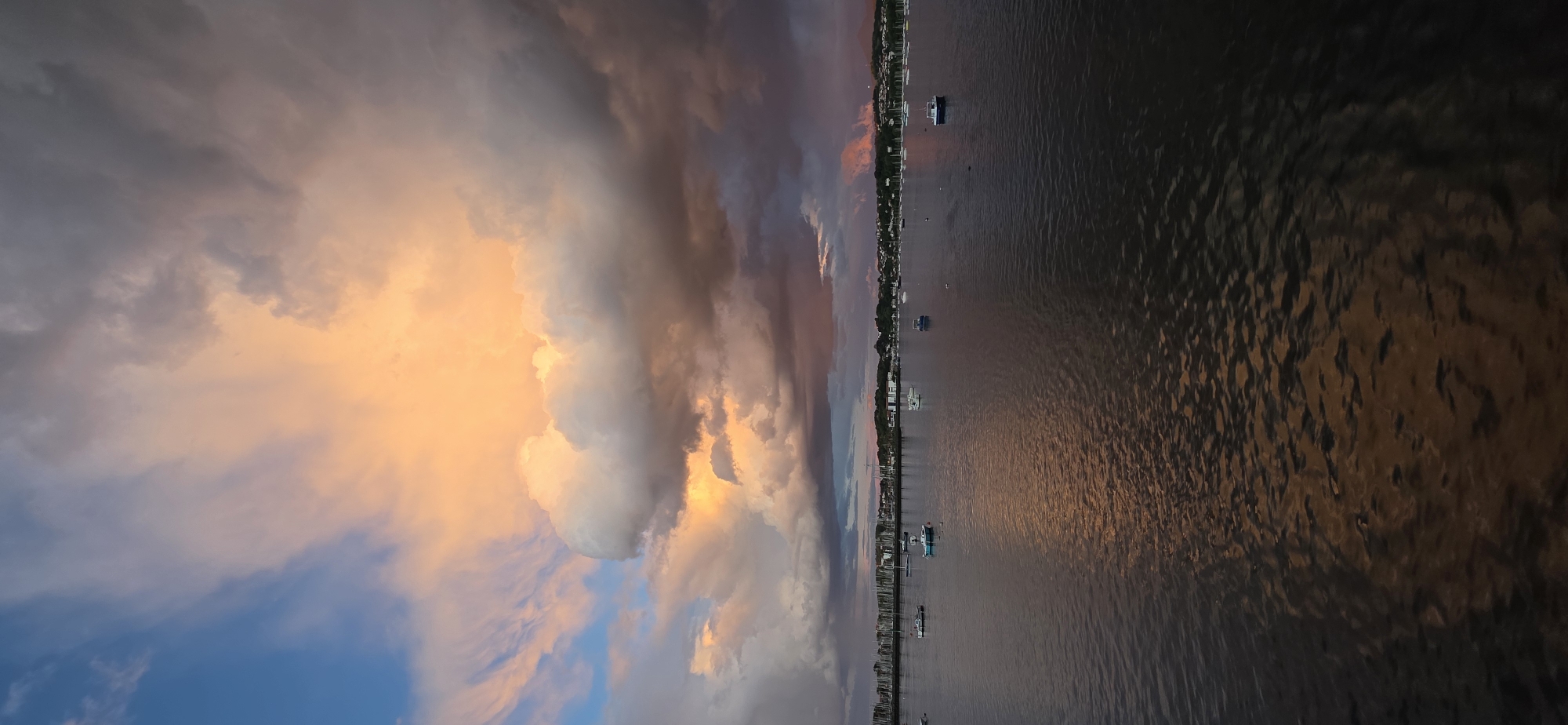 The image captures a sunset over the water in Hondarribia, Spain. The sky is filled with colorful clouds, with hues of orange, gray, and blue. A shoreline runs horizontally across the middle, where buildings and moored boats are visible. The water reflects the colors of the sky, creating a shimmering effect.