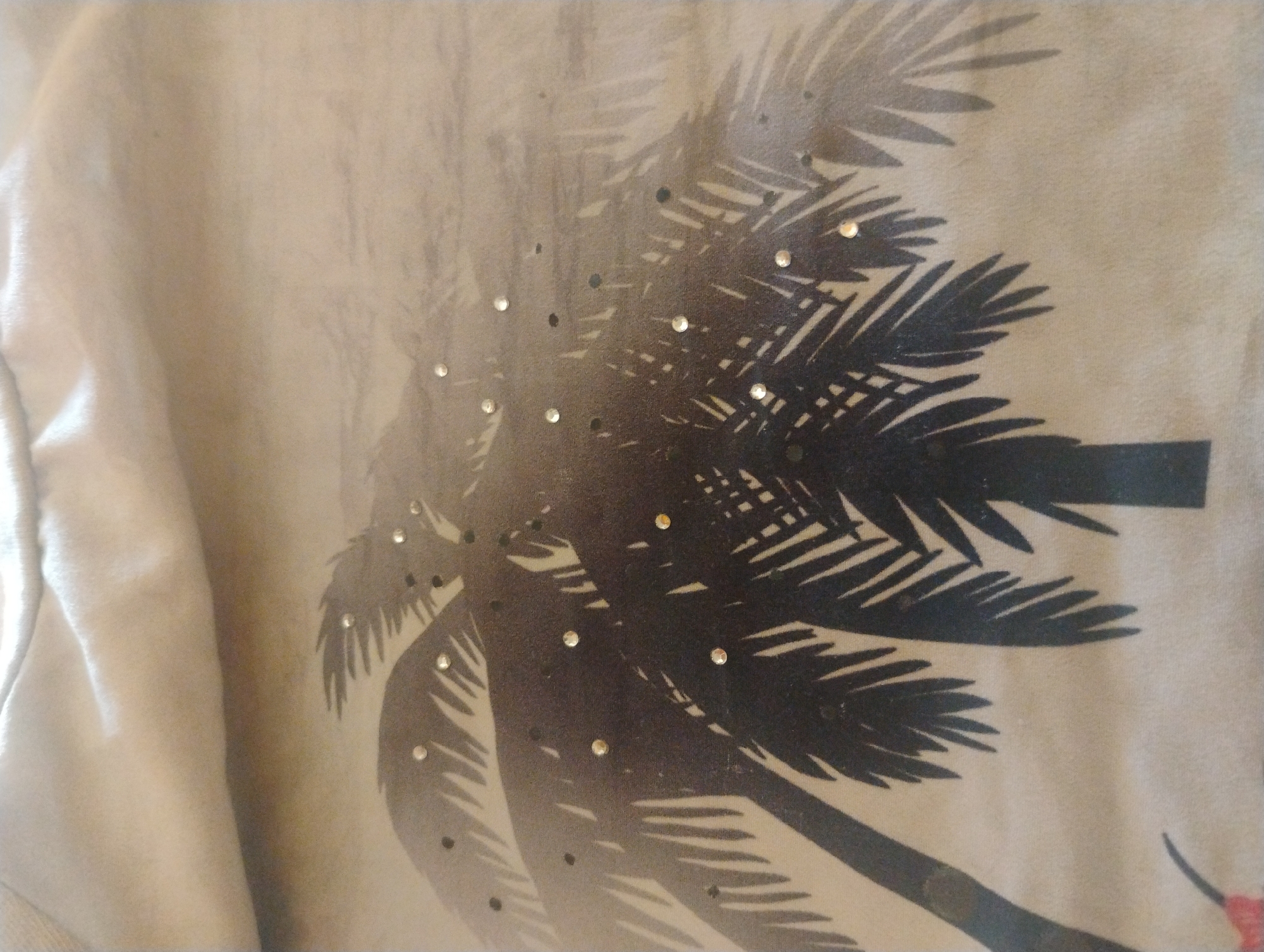 The image shows a close-up of a light-colored fabric, possibly a shirt, with a faded black palm tree silhouette printed on it. Small, reflective embellishments, likely rhinestones or sequins, are scattered across the palm fronds and in the background. The fabric appears to be slightly wrinkled, indicating it is a soft material. There is no indication of the location being in the city of Rumyantsevo, Bulgaria, as the image exclusively focuses on the fabric design. The scene is static and does not depict any people, actions, or interactions.