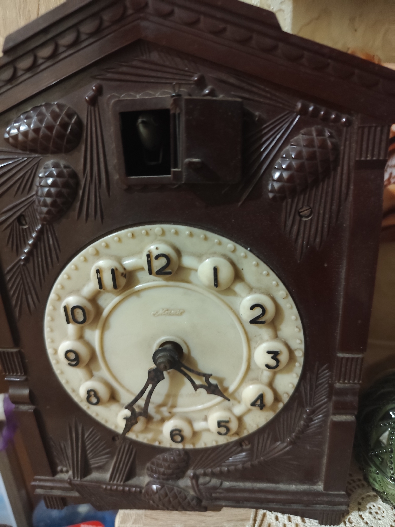 An antique cuckoo clock is the central focus of this image, displayed indoors against a wooden wall. The clock is dark brown and ornately carved with pinecones and foliage. The clock face is cream-colored with black numbers and hands, and a small, dark opening above the face where a cuckoo bird would emerge. The clock appears to be in a state of disrepair or inactivity. The surrounding environment suggests a domestic setting, with hints of other objects and textiles visible. The exact location within Montana, Bulgaria, is not identifiable from the image alone, but the overall impression is one of an aged, possibly inherited, timepiece.