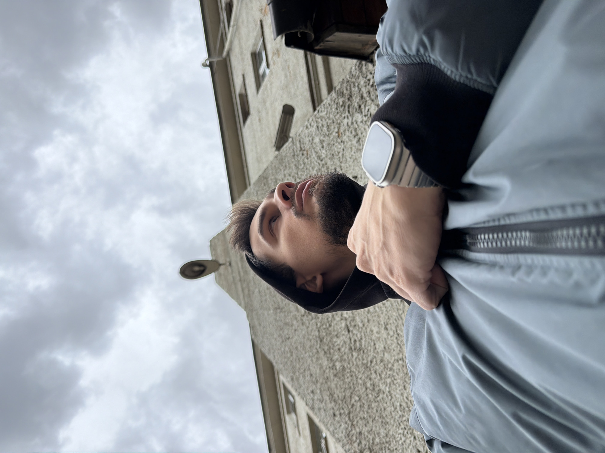 A low-angle shot of a man looking up, with a building and cloudy sky in the background. The man has brown hair and a beard, and he is wearing a hooded jacket and a smartwatch. The building is made of stone, and it has several windows. The street lamp and the building suggest the location could be Krasnoyarsk, Russia.
