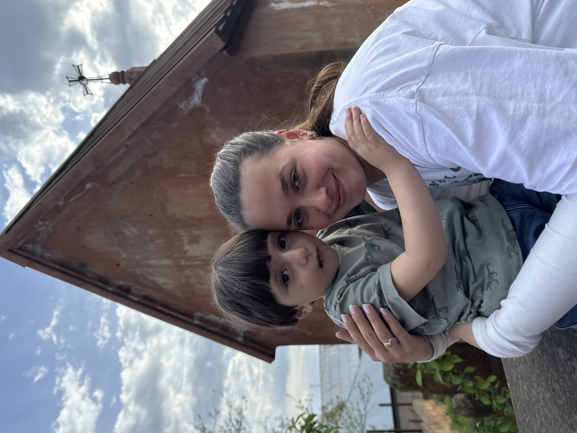 The image shows a woman and a young child standing in front of a building, presumably in Arinj, Armenia. The building has a cross on top. The woman is wearing a white t-shirt, the child is wearing a green t-shirt with a dinosaur pattern. The woman is hugging the child. The sky is visible in the background.