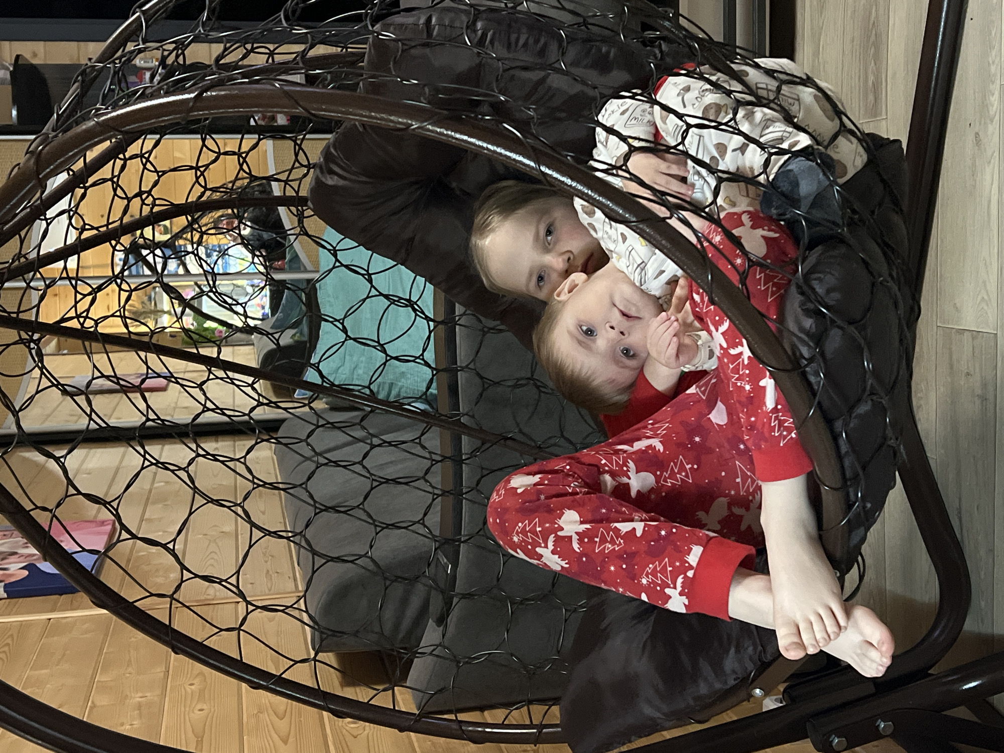 Two children are seated in a hanging pod chair indoors. One child is wearing red pajamas with white Christmas trees and reindeer, while the other child is wearing pajamas with a milk bottle print. The chair is dark brown with a mesh structure and has a dark brown cushion. The room has a wooden floor. A couch with throw pillows can be partially seen in the background. There is no indication of the location being Bol'shakino, Russia.