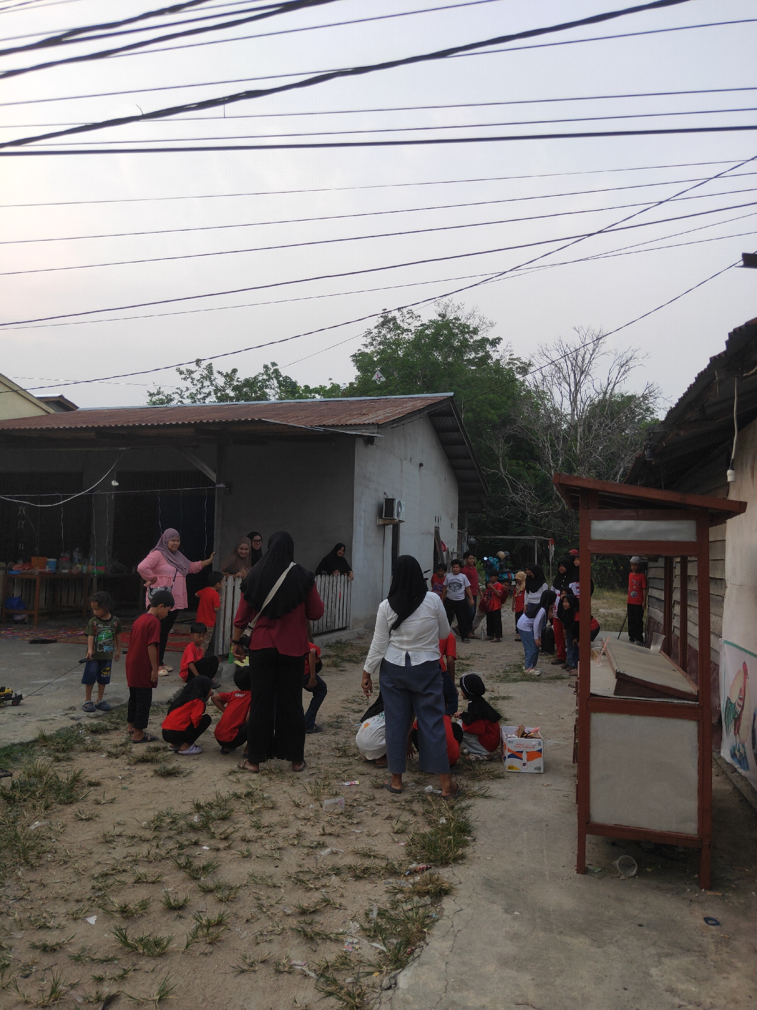 The image depicts an outdoor gathering of people, likely a community event, in a somewhat unkempt, rural or semi-rural setting. The environment appears to be a dusty, dirt and concrete ground with sparse patches of grass. Several buildings with corrugated metal roofs are visible, suggesting a residential or communal area. Overhead, multiple power lines crisscross the sky, indicating proximity to an electrical grid.
In the foreground and midground, a group of individuals, including adults and children, are engaged in various activities. Some are standing and talking, while others are seated or crouching. The participants are dressed in casual attire, with several children wearing red shirts. One woman in a pink headscarf appears to be speaking to a group. Another woman in a white shirt and blue pants is standing with her back to the camera. A wooden stall or kiosk with a red frame is prominent on the right side of the image. Scattered on the ground are a few plastic bags and some debris.
The lighting suggests it is either early morning or late afternoon, with a somewhat overcast sky. The overall impression is one of a casual, informal gathering in a modest setting. Without specific landmarks or identifiable signage, pinpointing the exact location within Sintang, Indonesia, is not possible solely based on this image. However, the scene is consistent with the appearance of many residential areas in the region.