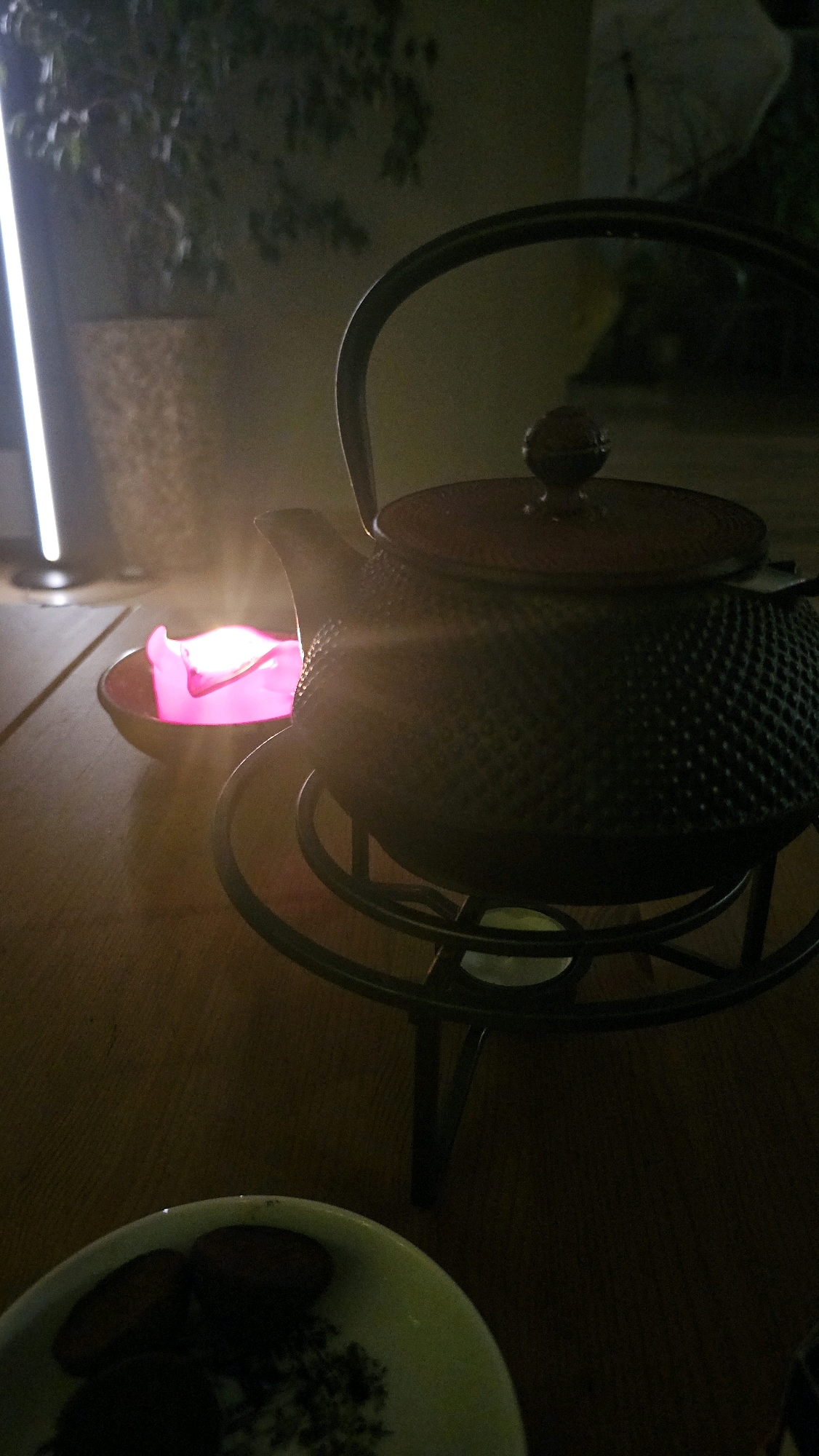 An indoor scene, likely a restaurant or home dining area in Cologne, Germany, is depicted. The lighting is dim, with a bright pink candle in a small bowl casting a warm glow on the left side of the frame. In the center, a dark, cast-iron teapot sits on a metal stand. The teapot has a textured surface and a rounded lid with a decorative knob. To the lower left, a portion of a white plate with a green rim is visible, containing several dark, circular objects, possibly food items like cookies or pastries. The background is blurred, showing dark greenery and a vertical light source, suggesting an outdoor or window view. The overall ambiance is intimate and dimly lit, with the candle and teapot as the primary focal points.