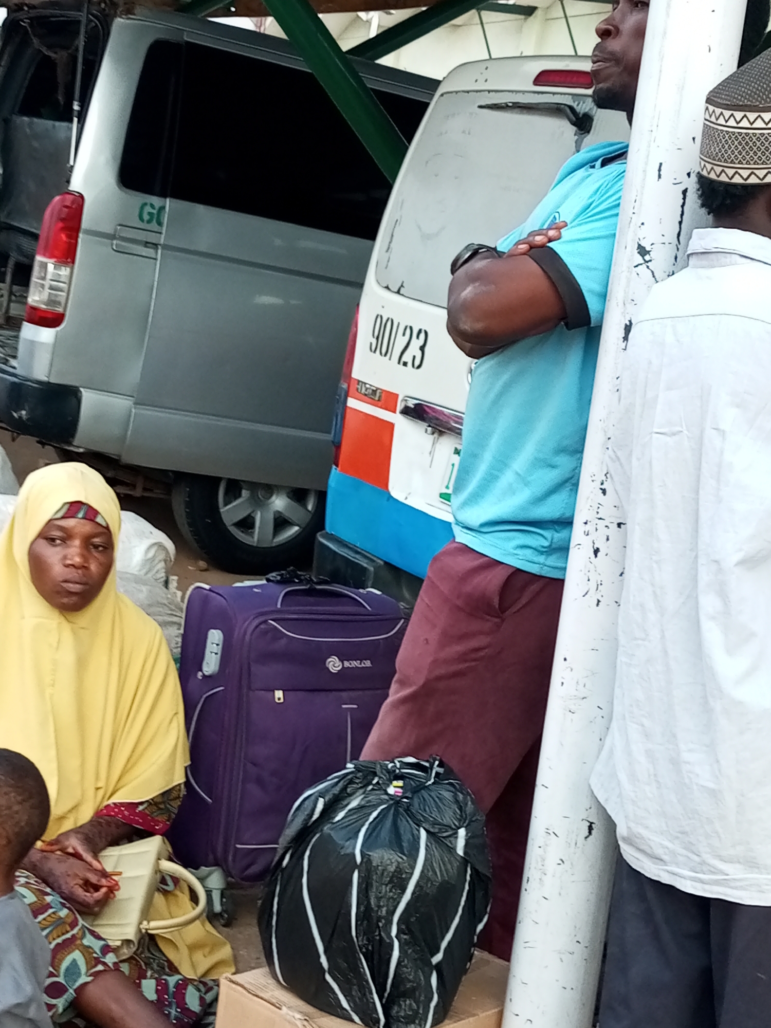 The image shows a street scene in Gombe, Nigeria. In the foreground, a woman wearing a yellow hijab and patterned clothing sits with her hands clasped, holding a small beige handbag. Next to her is a large, dark purple suitcase with the brand name "BONZAR" visible. To the right of the suitcase is a large black plastic bag filled with items, possibly trash or belongings. Behind the woman, a silver minivan with "GO" written on its side is parked. To the right of the minivan, a blue and white passenger van with the number "90/23" is visible. A man in a light blue t-shirt and dark red trousers stands with his arms crossed, leaning against a white pole. Another person, partially visible, is wearing a white shirt and a patterned cap, also standing near the pole. The environment appears to be outdoors, possibly a transportation hub or market area, with a covered structure overhead suggested by the green support beams.
