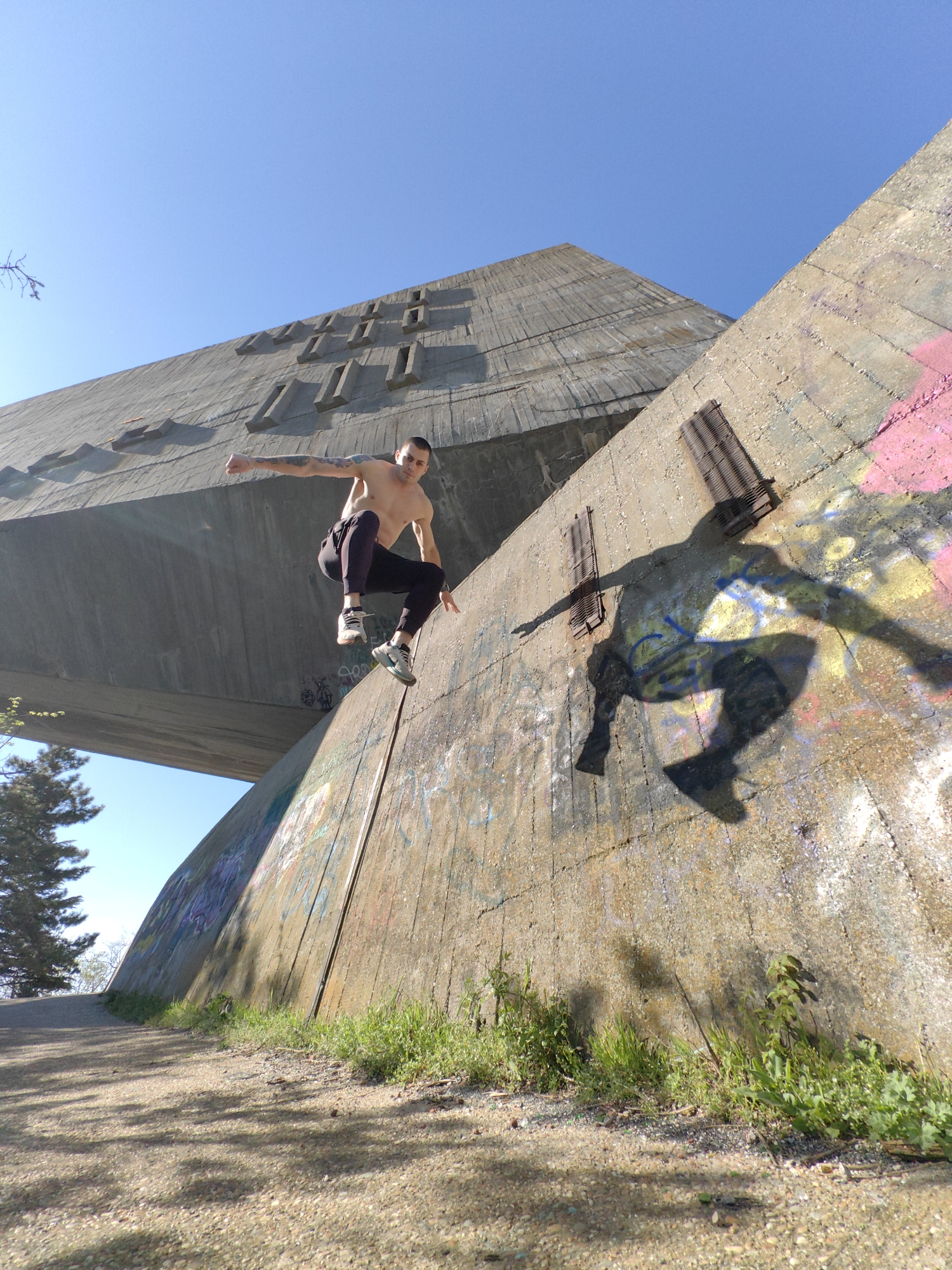 A man is jumping off a concrete structure in Varna, Bulgaria. He is fair-skinned, shirtless, and wearing dark pants and sneakers. His shadow is cast on the concrete wall behind him. The concrete structure is large and angular, with graffiti on the lower portions. A path runs along the base of the structure, with sparse green vegetation. The sky is clear and blue. The location is likely the Monument of the Bulgarian-Soviet Friendship in Varna.