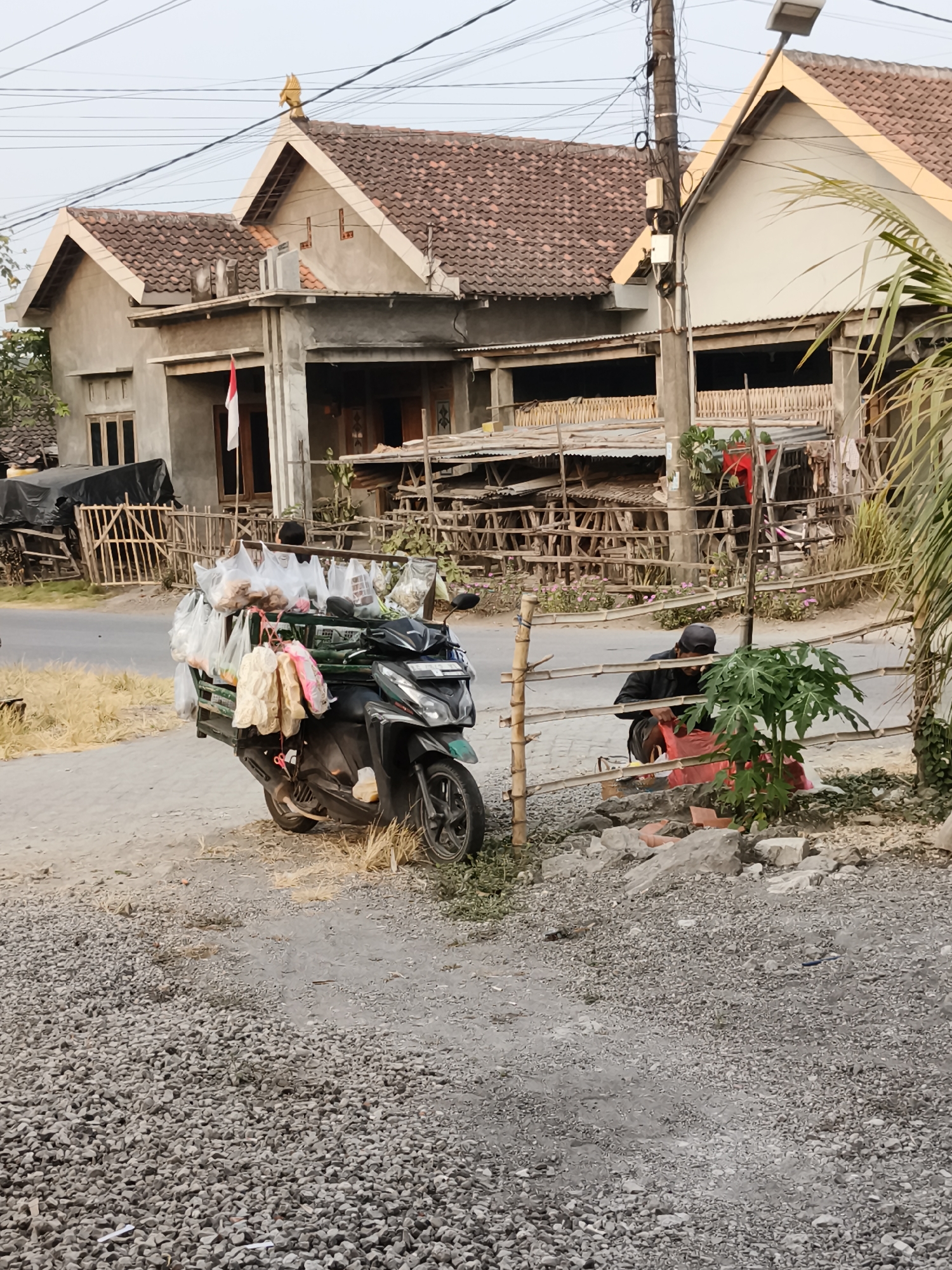 An outdoor, eye-level, medium shot captures a street scene in Pandean, Indonesia. The foreground is dominated by a gravelly surface, with a black scooter parked on the right side. The scooter is equipped with a rack containing numerous plastic bags filled with goods, appearing to be food items. A person, wearing a dark jacket and a baseball cap, is partially visible behind the scooter, tending to a red bag near a bamboo fence. The middle ground features a road and a bamboo fence bordering the properties. In the background, several houses with tiled roofs are visible, along with overhead electrical wires and a street lamp. A small Indonesian flag is displayed on the left-most house. The lighting suggests it is either early morning or late afternoon.