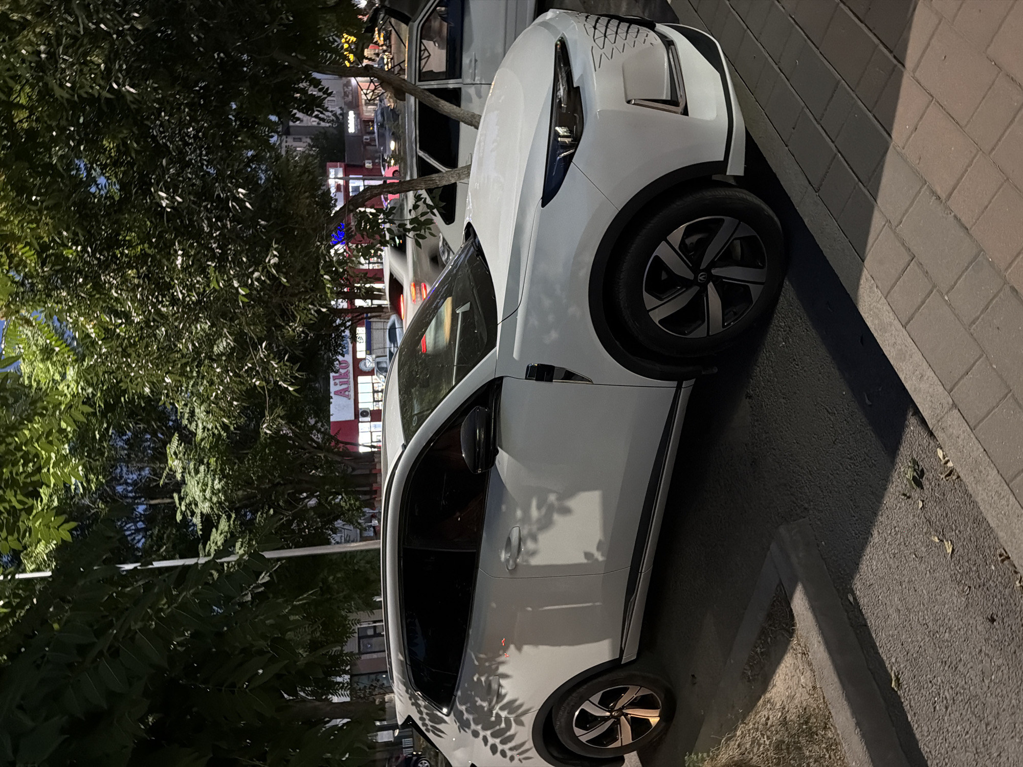 A white car is parked on the side of a street in Shymkent, Kazakhstan. The car is a modern model with sleek lines and distinctive wheel rims. Its front is angled towards the viewer, showing the headlights and a patterned design on the hood. To the left of the car, trees with dense foliage cast shadows on the vehicle. Behind the trees, buildings with illuminated windows and signs are visible, suggesting it is nighttime. One sign reads "AIKO" in red letters. The street is paved with asphalt, and a sidewalk made of rectangular bricks runs along the right side of the image. The overall scene depicts an urban setting at dusk or night.