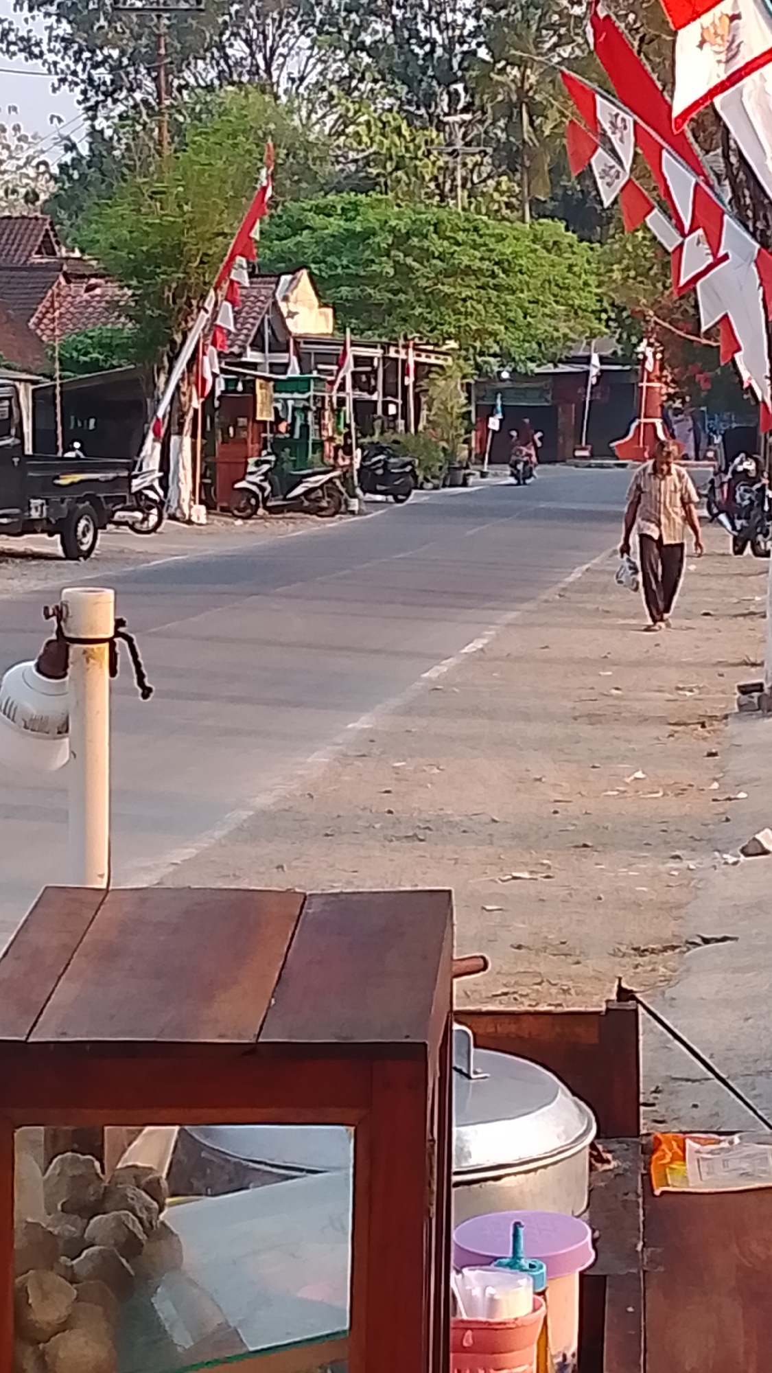 The image captures a street scene in Karanglangu, Indonesia, likely during daytime given the lighting. A paved road runs through the scene, with a sandy shoulder on the right. Red and white Indonesian flags are strung along poles on the left side of the road, suggesting a festive or patriotic occasion.
On the left side of the road, a black truck is parked. Several motorcycles are visible, parked or in motion on the road. A man in a striped shirt and dark pants is walking on the right side of the road, carrying a plastic bag in his right hand. In the foreground, a wooden food stall is partially visible, with a glass display case containing round food items. A large metal pot with a lid is also present behind the stall. The environment appears to be semi-rural or suburban, with trees and buildings lining the street in the background.