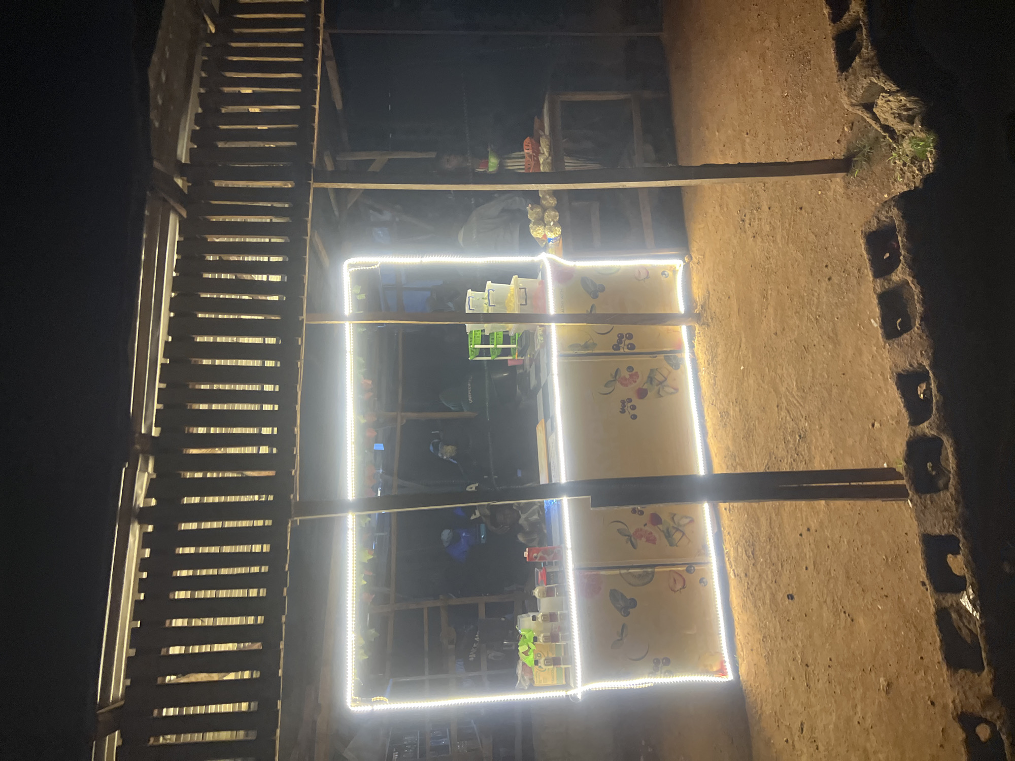 An outdoor food stall in Durumi, Nigeria, is brightly lit by a string of white LED lights outlining its front. The stall appears to be constructed of wood and corrugated metal roofing. The counter area displays various items, including bottles of liquids, what appear to be food containers, and a juice box. In the background, several individuals are visible inside the stall, some appearing to be engaged in food preparation or service. The ground in front of the stall is dirt, and a low brick structure is visible in the foreground. The overall scene suggests a bustling nighttime operation.