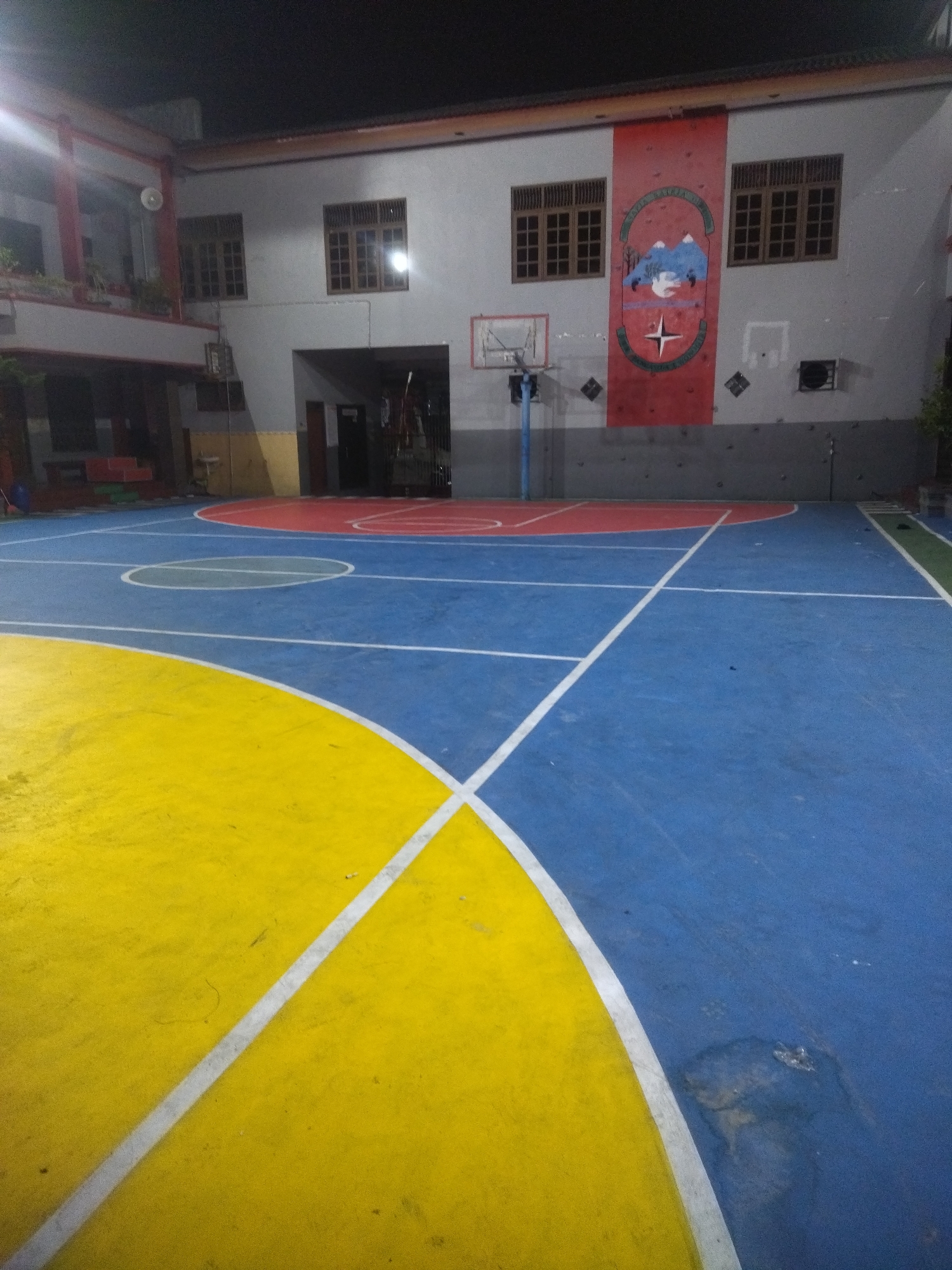 The image depicts an outdoor basketball court at night, likely in Giritirto, Indonesia, given the context. The court features distinct colored sections: a large area of blue with white lines marking court boundaries and a smaller section of yellow. A red semi-circle is visible near a basketball hoop, which is mounted on a pole. Behind the court, a building with multiple windows and a red banner displaying a crest is visible. The lighting suggests it is evening or night. There are no people or animals present in the scene.