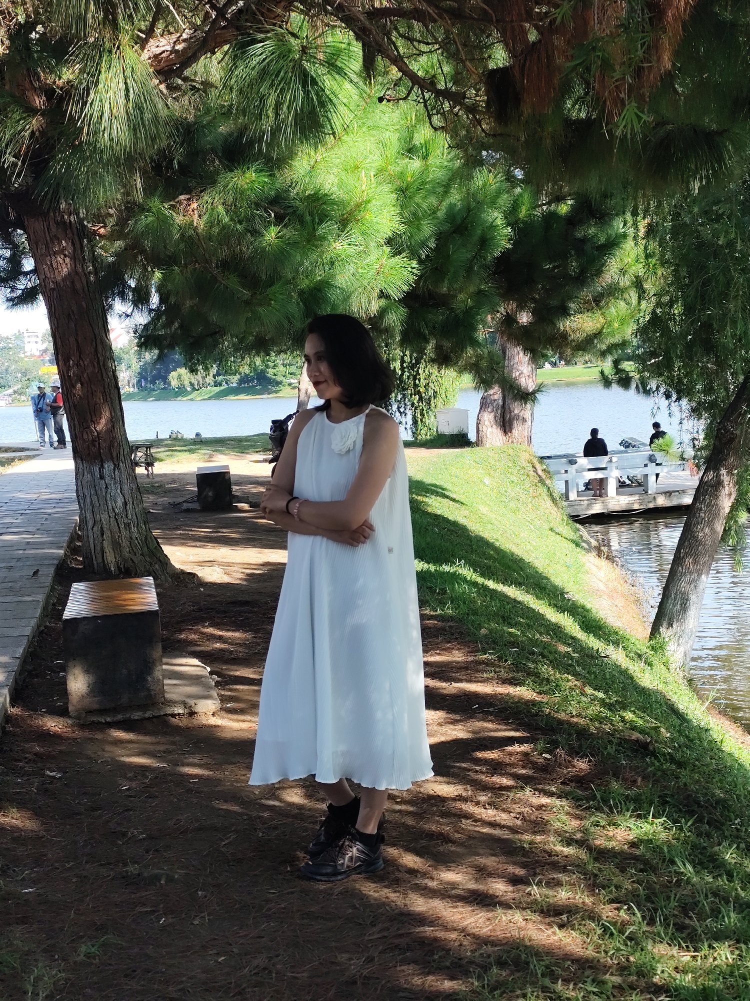 A woman in a white dress stands with her arms crossed under the shade of pine trees next to Xuan Huong Lake in Da Lat, Vietnam. The woman, with dark hair and fair skin, is wearing a knee-length, sleeveless white dress with pleats and a floral embellishment on the left shoulder. She is also wearing dark sneakers. To her left, a wooden pathway runs alongside the lake, and two people, a man and a woman, walk in the distance. To her right, a grassy slope leads down to the water, where a small wooden pier extends into the lake, with two individuals standing on it. The lake reflects the surrounding greenery and the sky, which appears partly cloudy. The ground beneath the trees is covered with pine needles and dry leaves, with shadows cast by the branches. In the background, buildings can be seen on the distant hills.