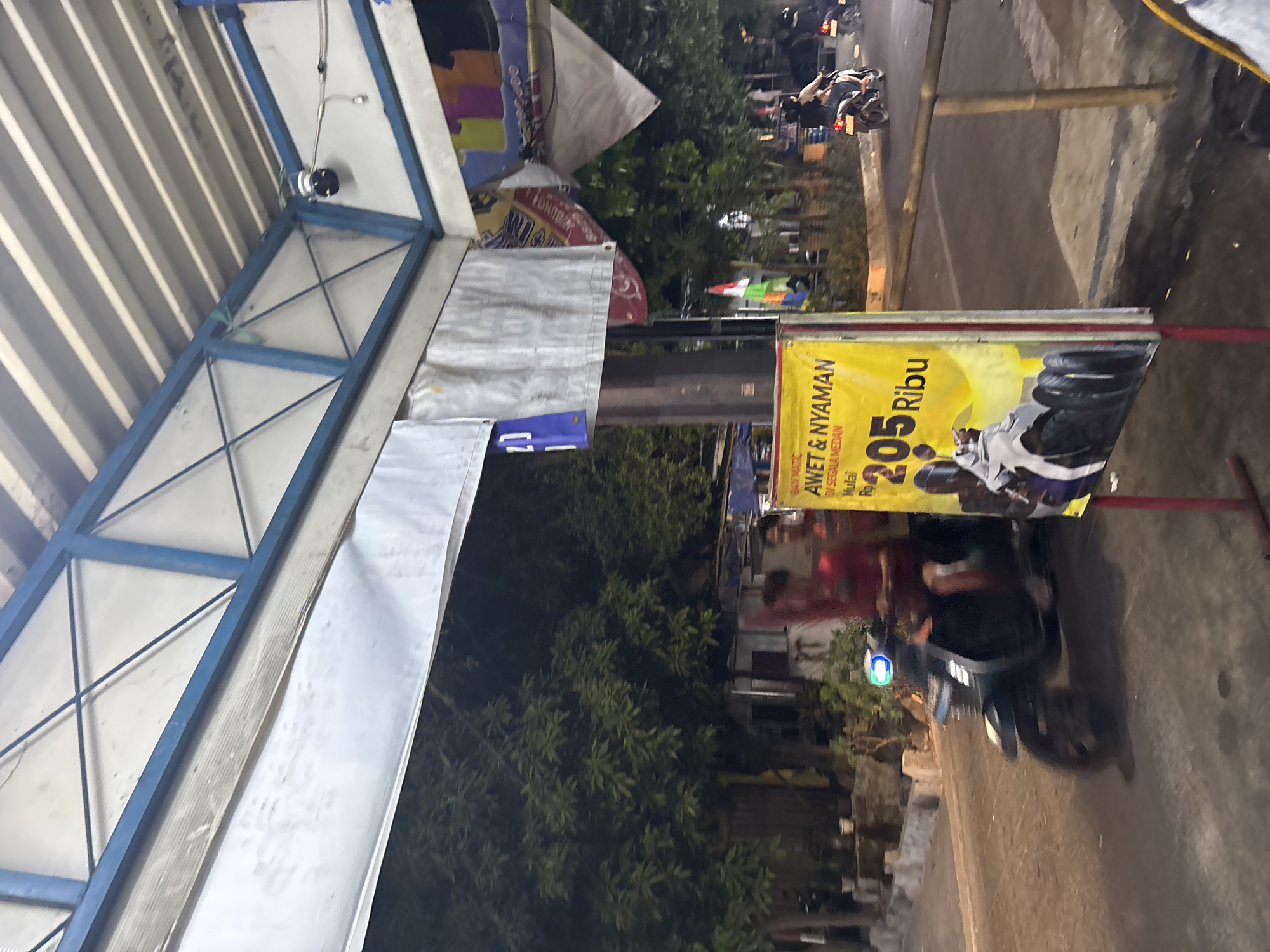 In a dimly lit outdoor scene in North Jakarta, Indonesia, a man wearing a red shirt rides a dark-colored scooter with a blue light on the front. He is moving from left to right across the frame. In the foreground, a yellow advertisement sign displays text in Indonesian, including the price "205 Ribu" which translates to "205 Thousand". The sign also features an image of several tires. Behind the scooter and the sign, a densely vegetated area with trees is visible, suggesting a suburban or slightly rural edge within the city. Above, a blue and white structure with a corrugated metal roof, possibly a shelter or canopy, is present. The overall lighting suggests it is evening or night, with artificial lights illuminating parts of the scene.