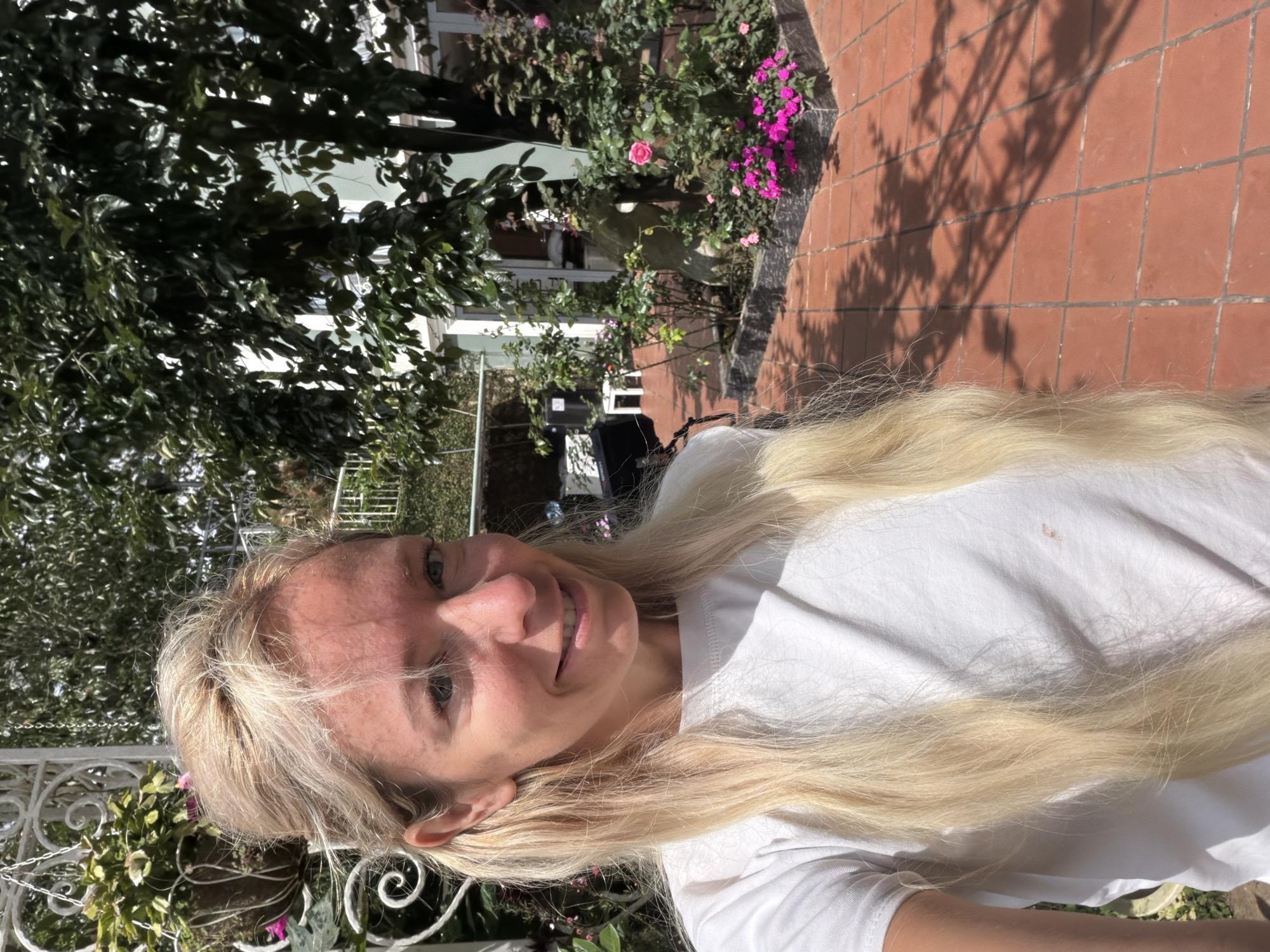 A woman with blonde hair and a white t-shirt is standing in a courtyard with red tile flooring and a garden. She is smiling and looking at the camera. Behind her, there is a tree with dark green leaves, a bush with pink roses, and other plants. There is also a white structure with hanging plants to her left. In the background, a building with a light green exterior is visible. The scene appears to be outdoors, in a sunny location, possibly in the city of Xuân Trường - Đà Lạt, Vietnam.