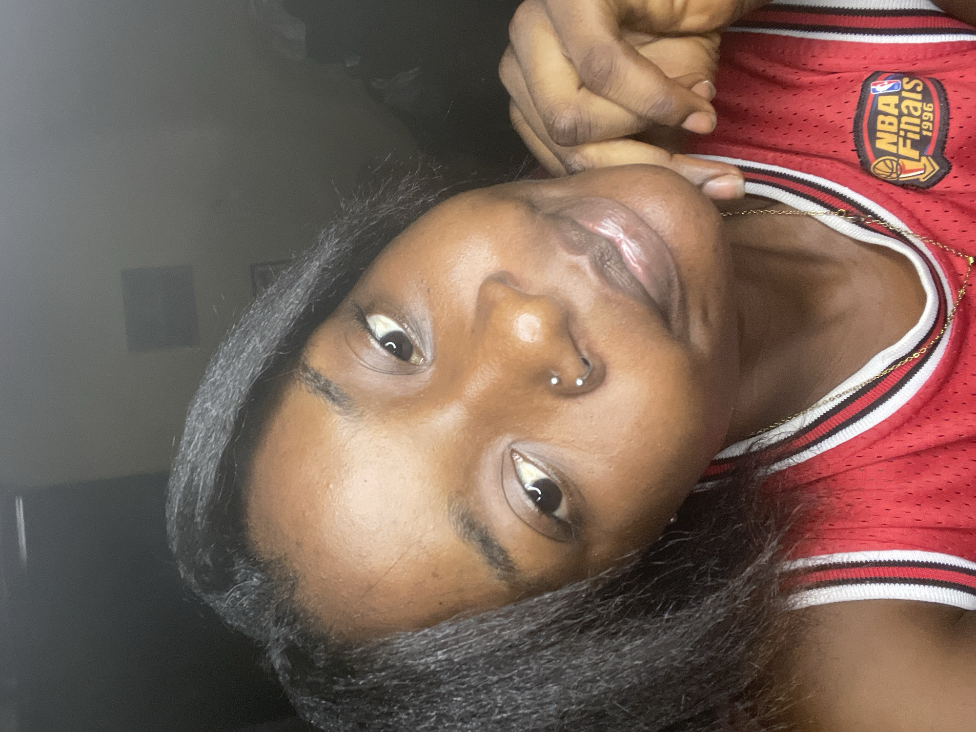 A close-up, medium shot shows a young Black woman with dark hair and brown skin. She is wearing a red basketball jersey with white and black trim, and a gold chain necklace. Her right hand is resting on her chin, and she is looking at the camera with a slight smile. Her eyes are open, and her lips are slightly parted. She has a nose ring with two small balls. The background is dark and out of focus, with no discernible details about the environment.