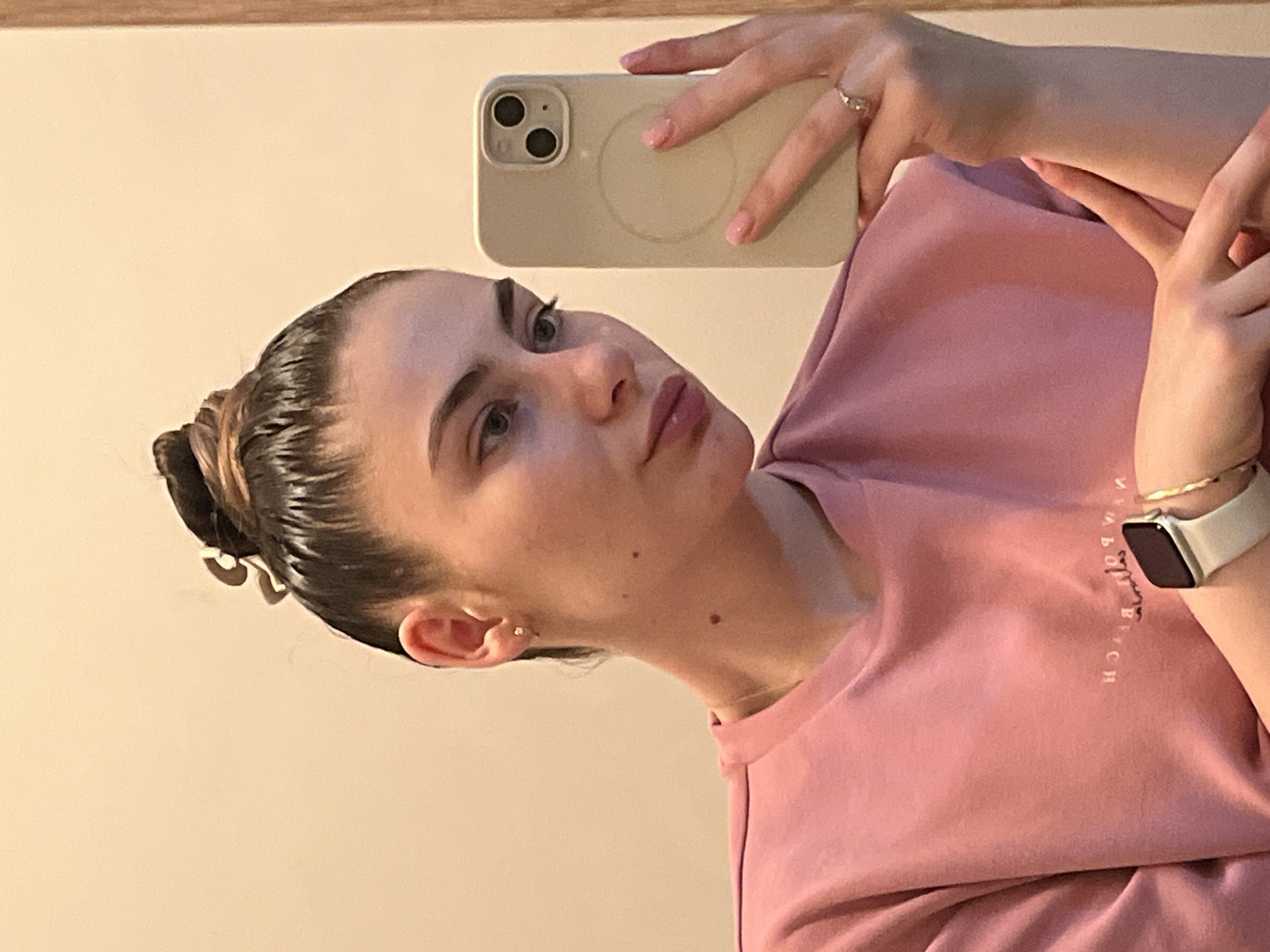 A young woman is captured in a mirror selfie, holding a light-colored smartphone with a dual-camera system and a visible magnetic ring on its case. She is looking towards the camera's reflection with a neutral expression, her lips slightly parted. Her hair is slicked back into a neat bun, revealing small earrings. She is wearing a plain pink long-sleeved top, which has light-colored, reversed text visible on the front, appearing to spell "NEW YORK". A silver-toned ring is visible on her right ring finger, and her fingernails are painted a light pink. On her left wrist, she wears a smartwatch with a light-colored band and a dark screen. The background is a plain, light-colored wall, possibly part of an indoor setting, with a hint of a wooden frame or ledge at the top edge of the mirror.
