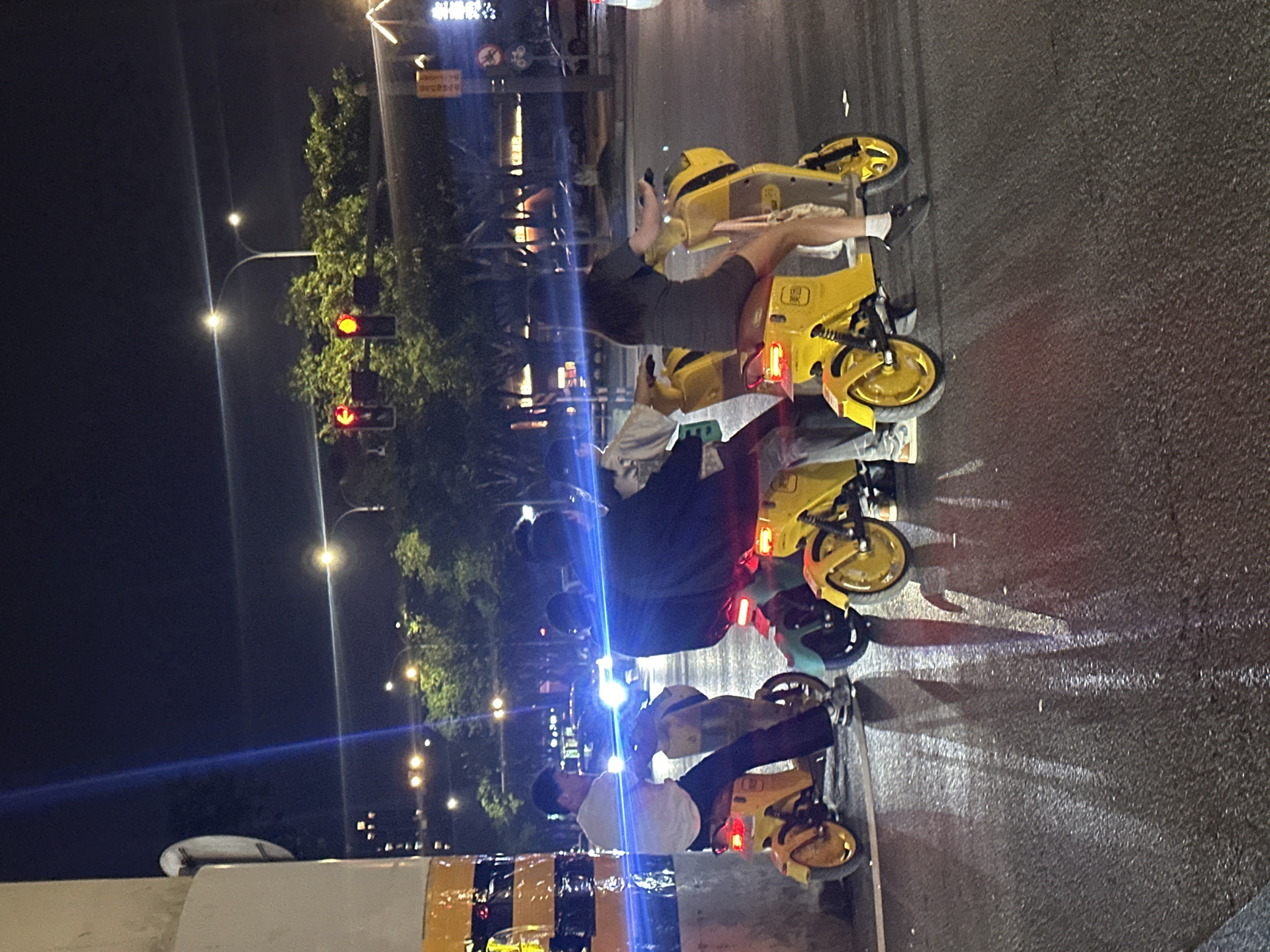 In the city of Chongqing High-tech Zone, China, at night, several individuals are riding shared yellow electric scooters on a wet asphalt road. The road reflects the lights of the scooters, traffic signals, and streetlights. In the foreground, three yellow scooters are visible, each with a rider. The leftmost scooter has a person wearing a white t-shirt and dark pants. The middle scooter has two people, one in a dark jacket and the other in a lighter-colored top. The rightmost scooter has a person wearing a dark dress. Traffic lights are illuminated red in the background, suggesting the riders are stopped. Trees and buildings are visible in the distance, along with streetlights. A large, striped pole is present on the left side of the image.