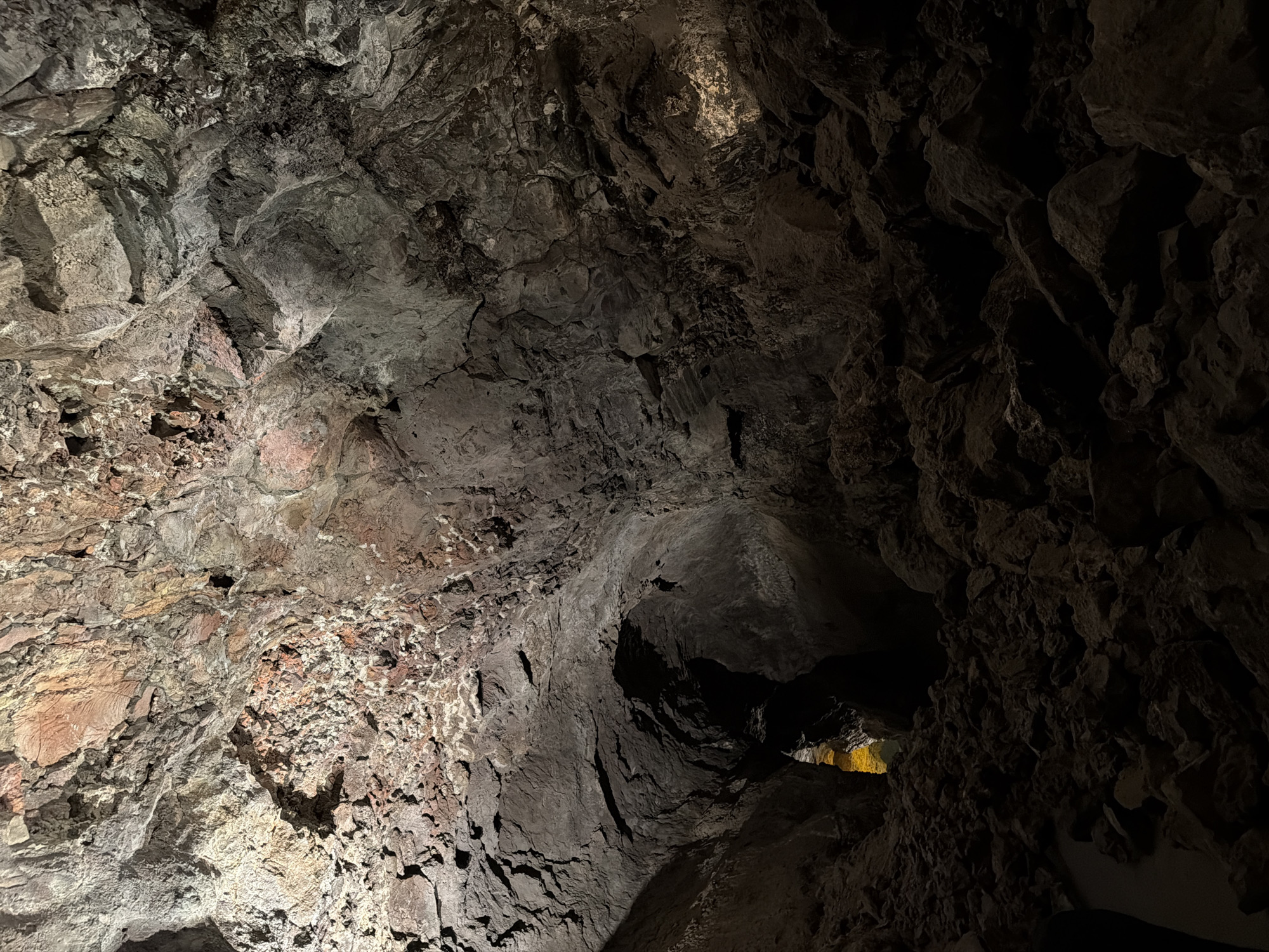 The image depicts the interior of the Cueva de los Verdes, a lava tube located in Haría, Lanzarote, Spain. The scene is largely dominated by the cave's rocky, textured walls, illuminated unevenly by artificial lighting, creating areas of bright exposure and deep shadow. The rock varies in color from reddish-brown to dark gray. In the distance, a small opening glows with artificial light. No people or other specific subjects are visible in the frame.