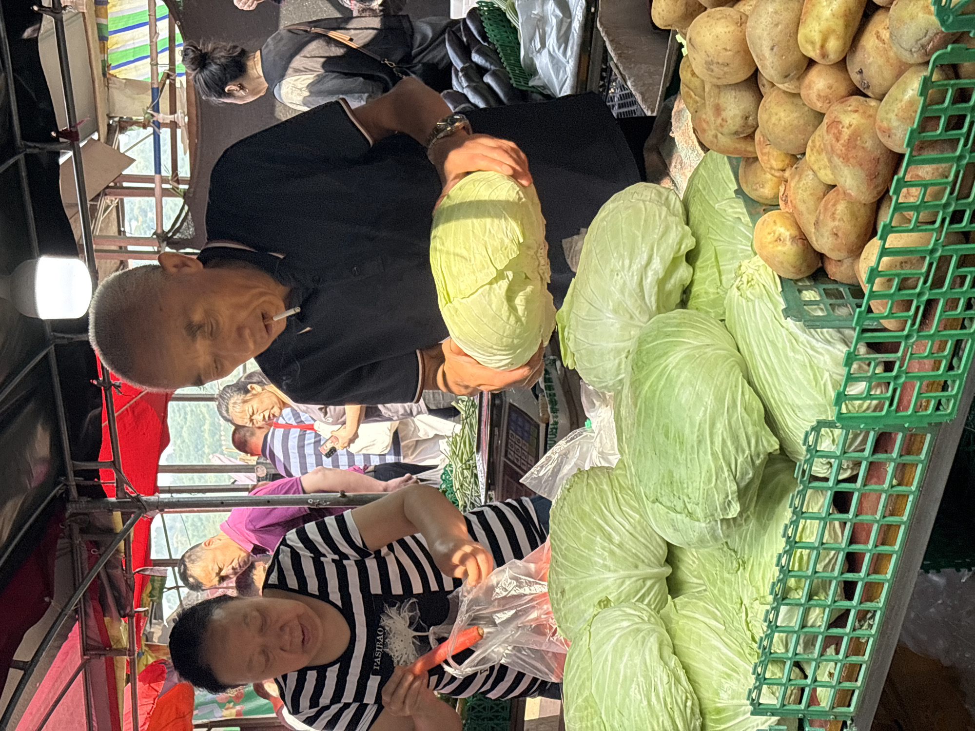 A street market scene in Jiangjin, China. A man in a black shirt with a cigarette in his mouth holds a head of cabbage. Another person in a black and white striped shirt is putting a carrot into a plastic bag. A pile of cabbages are displayed to the right, beside a green plastic crate filled with potatoes. Other people are visible in the background. A metal structure is visible overhead, possibly a part of the market stalls.