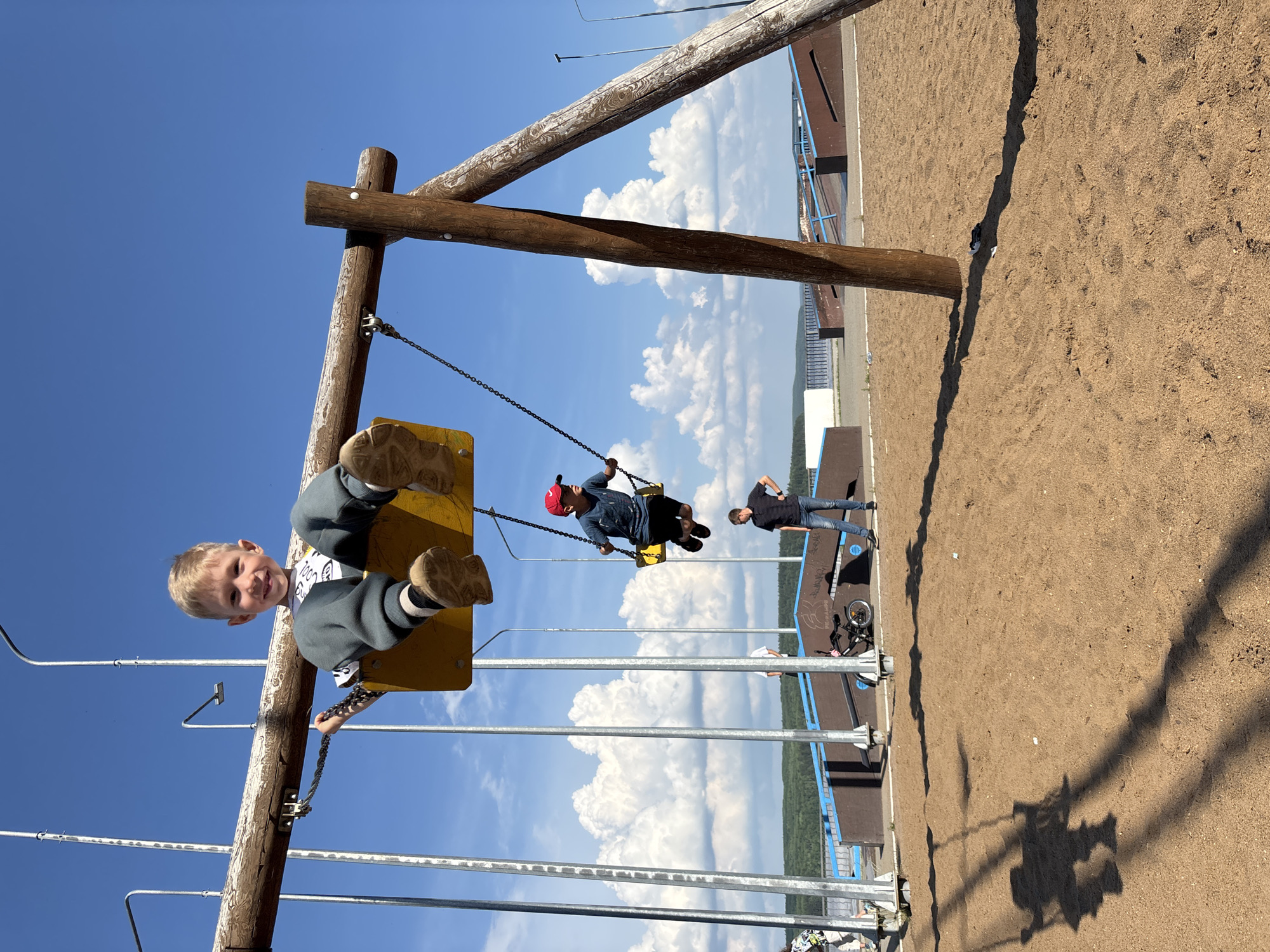 A medium shot captures a sunny day at a playground near the Kama River in Chaikovsky, Russia. In the foreground, a young boy with blonde hair is happily swinging on a wooden swing set. His legs are extended, and he is smiling towards the camera. Behind him, another boy in a blue shirt and red baseball cap is also on a swing. Further back, a third boy in a striped shirt stands on the sand, observing. The playground area features a sand pit for the swings and a skate park in the background with ramps. Tall metal poles and a large wooden beam support the swing set. The sky is a vibrant blue with scattered white clouds, and lush green trees line the horizon, suggesting a natural setting adjacent to the urban environment.