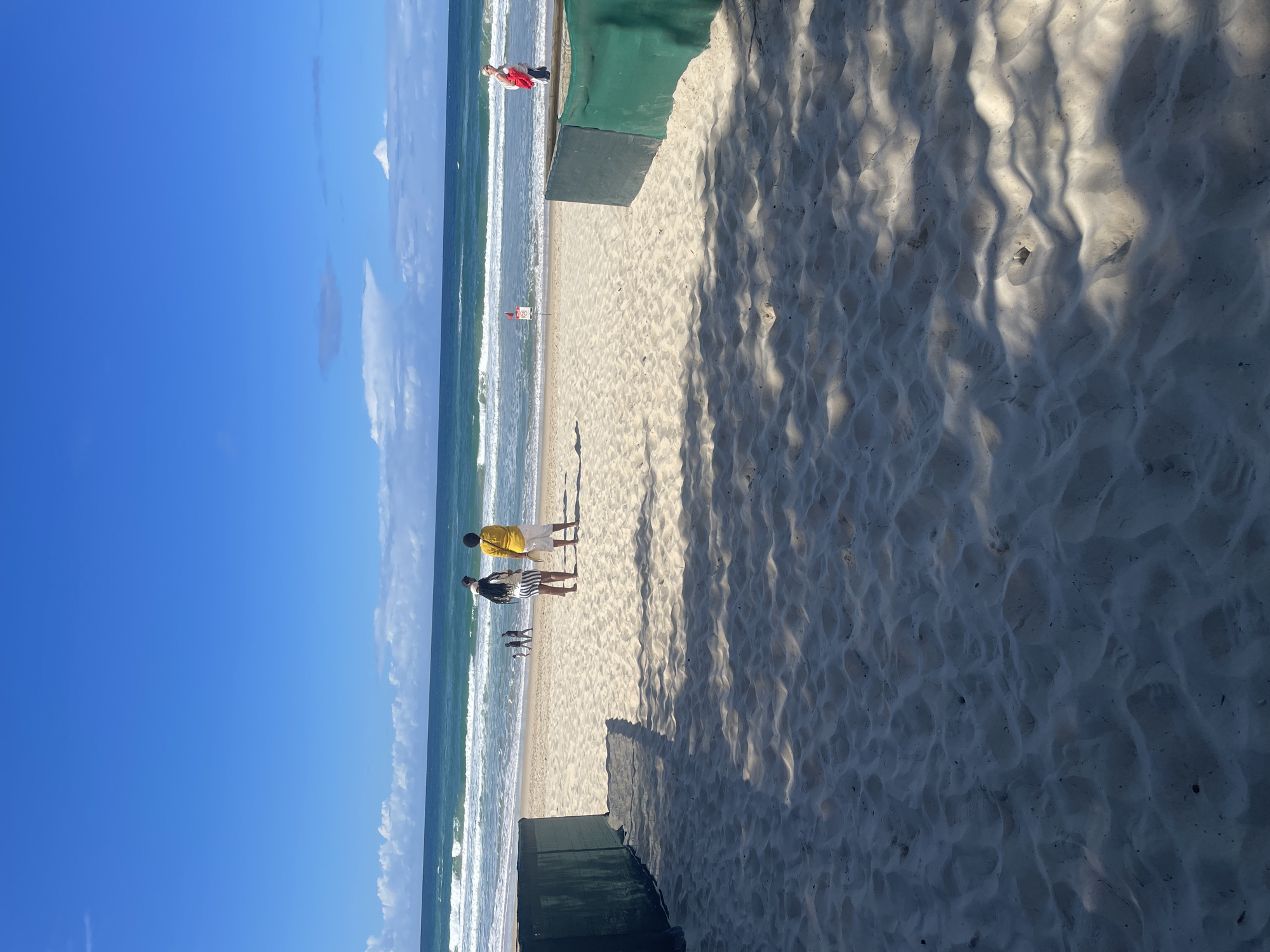 This image depicts a sunny day at a beach on the Gold Coast, Australia. The foreground features a sandy path leading towards the ocean, bordered by green netting on either side, casting dappled shadows on the sand. In the mid-ground, several people are visible on the beach. Two individuals stand facing the ocean, one wearing a yellow shirt and white shorts, and the other a black and white striped skirt and a dark top. Further back, a few other people are walking along the shoreline. The ocean exhibits moderate waves, with white surf breaking near the shore. The sky is a vibrant blue with scattered white clouds. A red flag and a sign are planted on the beach near the water's edge. A person in a white top and red jacket is walking further down the beach. The overall scene is that of a typical beach day with people enjoying the coastal environment.