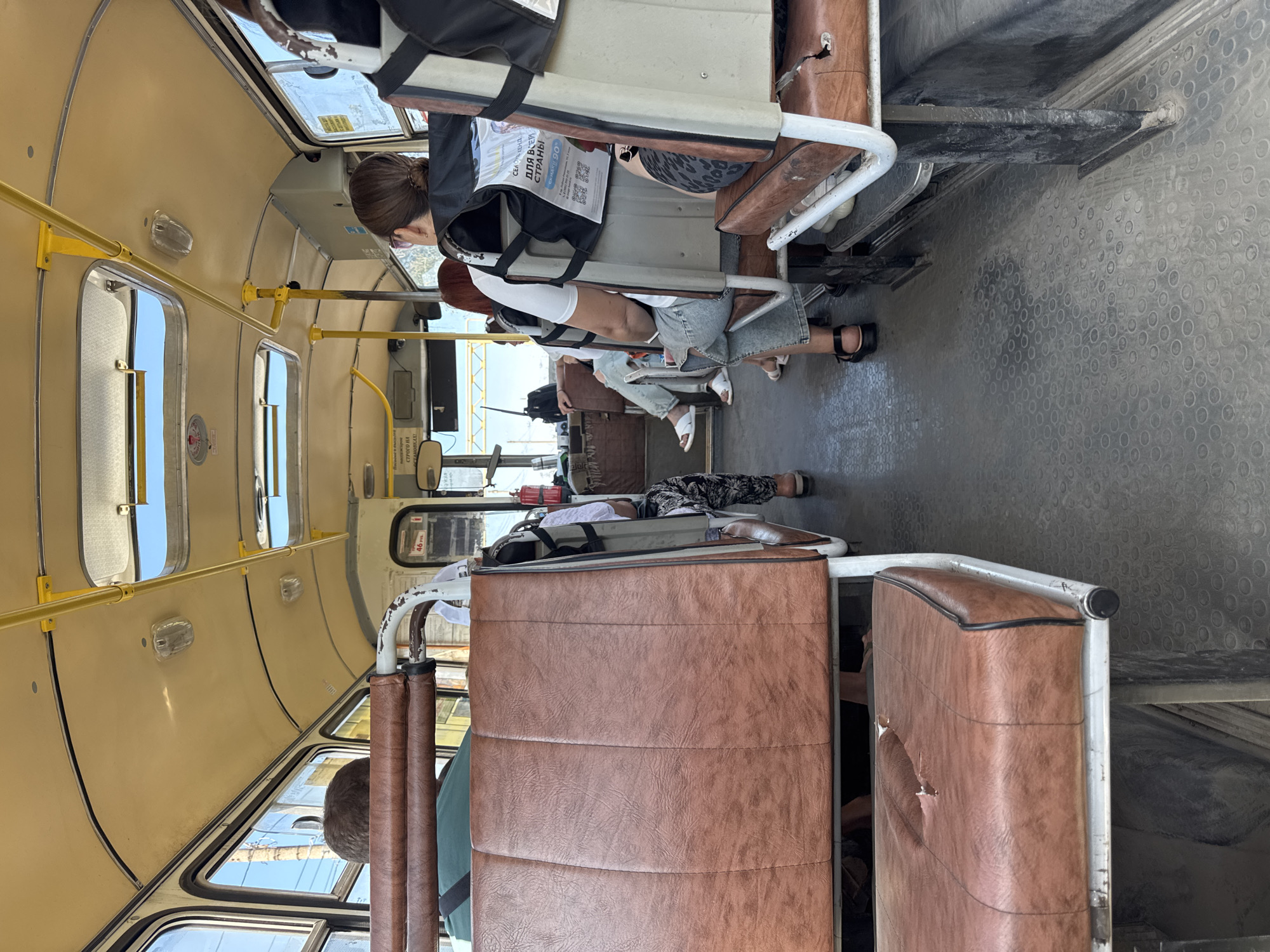 The image captures the interior of a bus, likely a public transportation vehicle, with passengers seated. The seats are upholstered in a brown, faux-leather material, and some show signs of wear, including a tear on the front-left seat. The ceiling is a pale yellow color, with visible yellow support poles and overhead lighting fixtures. Several passengers are visible from behind or the side, dressed in casual clothing. Some are looking forward, while others appear to be looking around or towards the front of the bus. On the back of one seat, a flyer or poster with text and a QR code is attached. The floor of the bus is a dark, textured material with a circular pattern. The overall impression is of a functional, though somewhat aged, mode of public transport. The scene is indoors, within the confines of the bus, with natural light entering through the windows. There is no specific indication of the location within Shakhty City District, Russia, other than the general context of a bus.