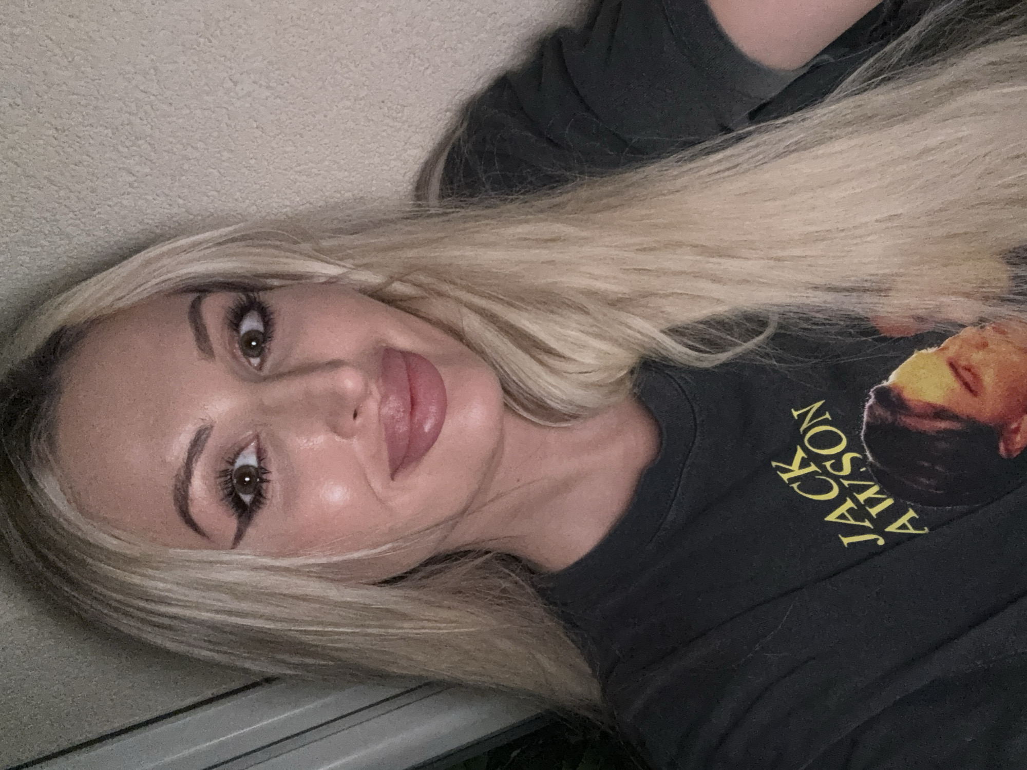 The image shows a selfie of a woman with long blonde hair. She is wearing a dark t-shirt with yellow text that reads "JACK AWSON" and a picture of a man. The background appears to be a wall. The woman has on makeup, including winged eyeliner and lip filler. Her expression is a slight smile. The lighting suggests the photo was taken indoors.