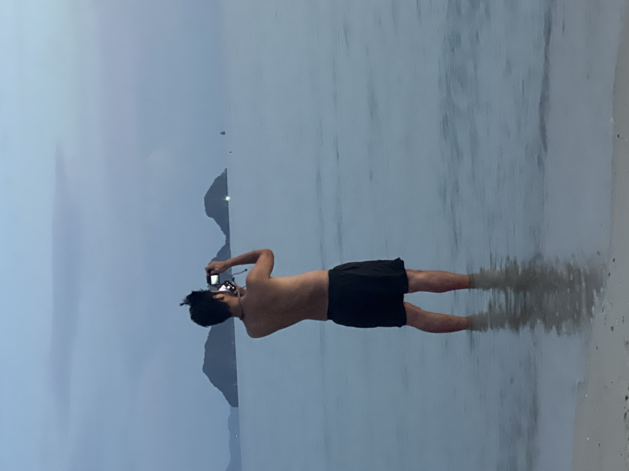 An individual stands waist-deep in the ocean at dusk, holding a camera to take a photograph. The person is shirtless, wearing black shorts. In the background, several islands are visible on the horizon, as well as a small boat. The water is calm, with gentle waves lapping at the shore. The sky is a muted blue, indicating the transition from day to night. The scene is peaceful and serene, capturing a moment of quiet contemplation by the sea. The location is the coast of Ko Pha-ngan District, Thailand.