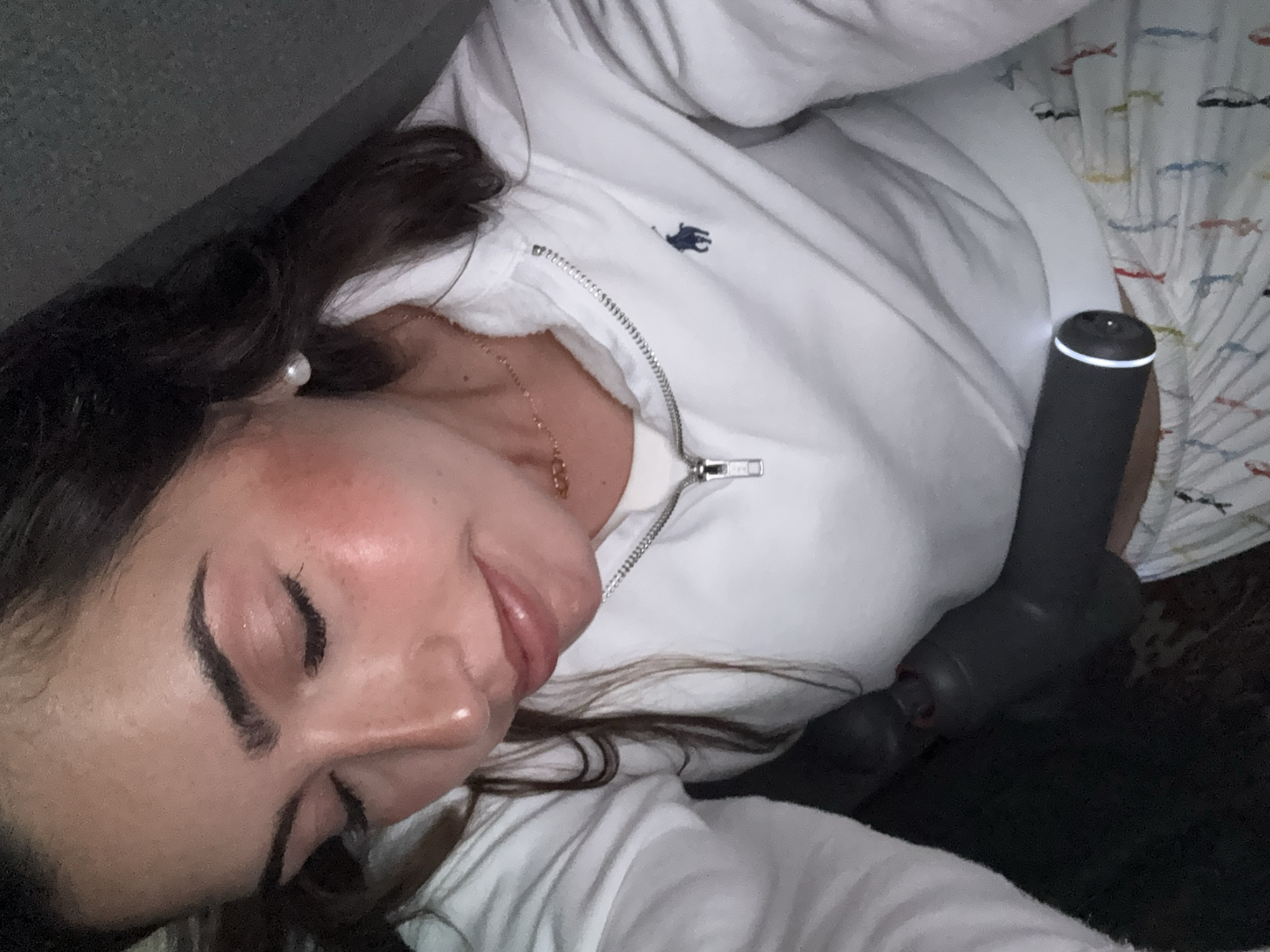 A woman lies in a resting position indoors. She has dark hair and is wearing a white zip-up jacket with a blue logo. A massage gun is situated near her body. The environment appears to be a bedroom, with patterned bedding visible in the background. There is no information to suggest that this image was taken in San Bernardino Tlaxcalancingo, Mexico.