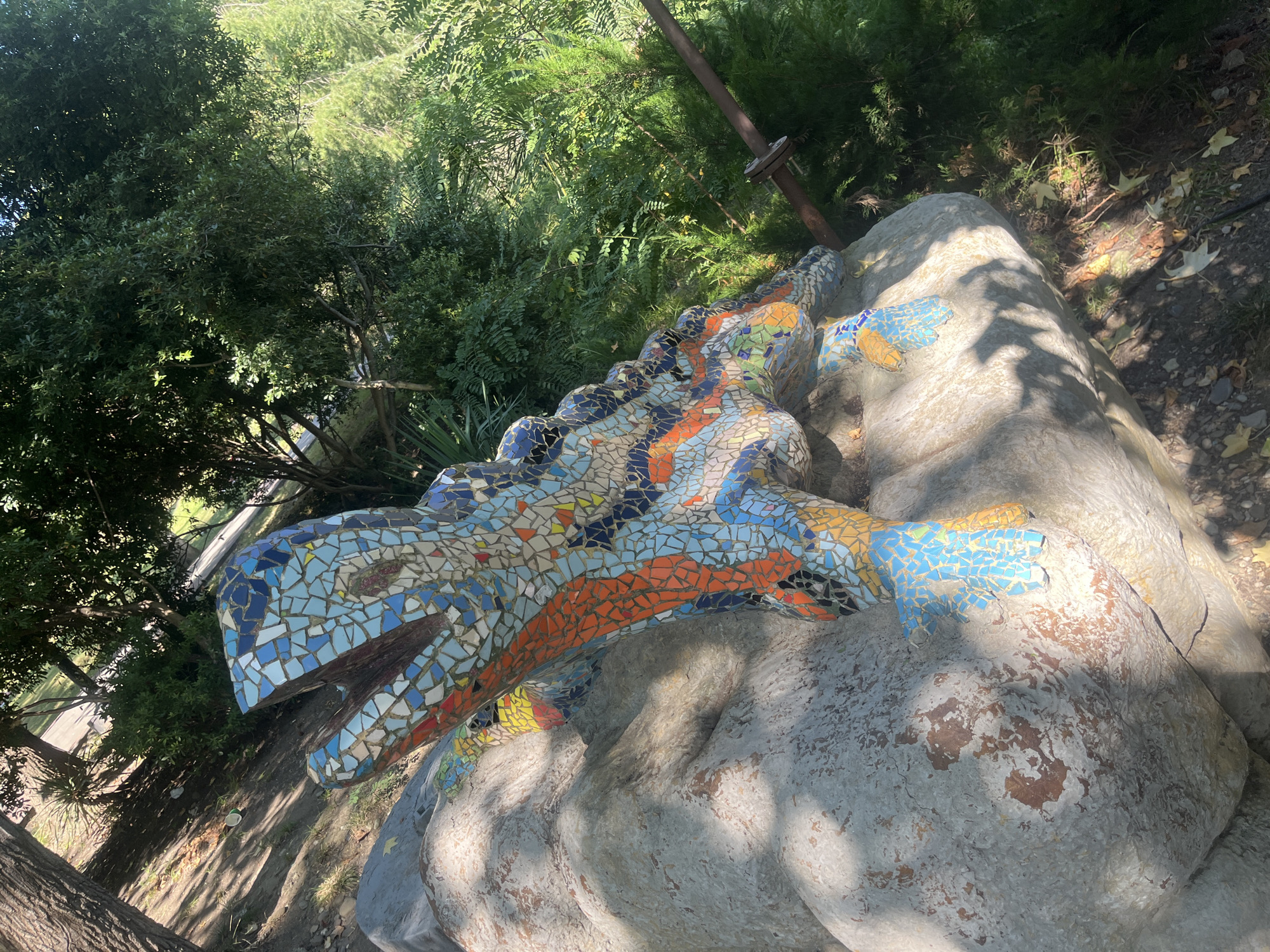 A mosaic sculpture of a lizard is perched on a large, light-colored rock. The lizard is covered in small, colorful mosaic tiles in shades of blue, orange, yellow, and white, with some black tiles outlining features. The sculpture is positioned outdoors, with lush green trees and foliage in the background. Sunlight filters through the trees, casting shadows on the rock and the lizard. The ground is covered with dirt, some fallen leaves, and sparse grass. This scene is located in the Dendrarium Botanical Garden in Sochi, Russia.