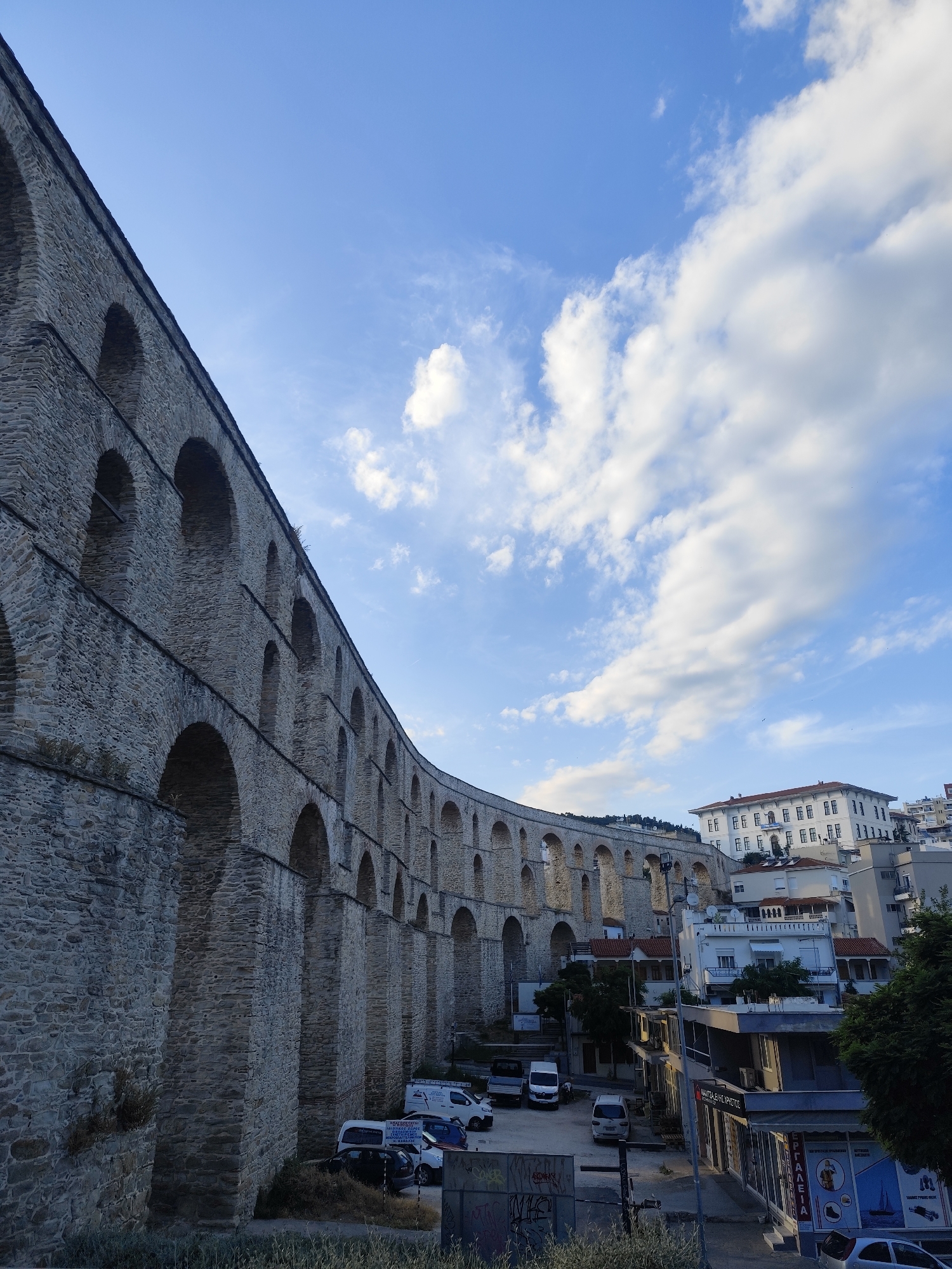 This image depicts the Kamares Aqueduct in Kavala, Greece, under a bright blue sky with scattered white clouds. The aqueduct, a massive stone structure with multiple arches, dominates the left side of the frame, curving towards the distance. Its textured stone construction is clearly visible. Below and to the right of the aqueduct, a street scene unfolds. Several buildings, including a large white building with many windows and a tiled roof, are situated on a hillside. Some smaller structures with balconies and tiled roofs are also present. A parking area is visible in front of some of these buildings, with various vehicles including vans and cars parked there. The overall impression is one of historical architecture integrated with a modern urban environment.