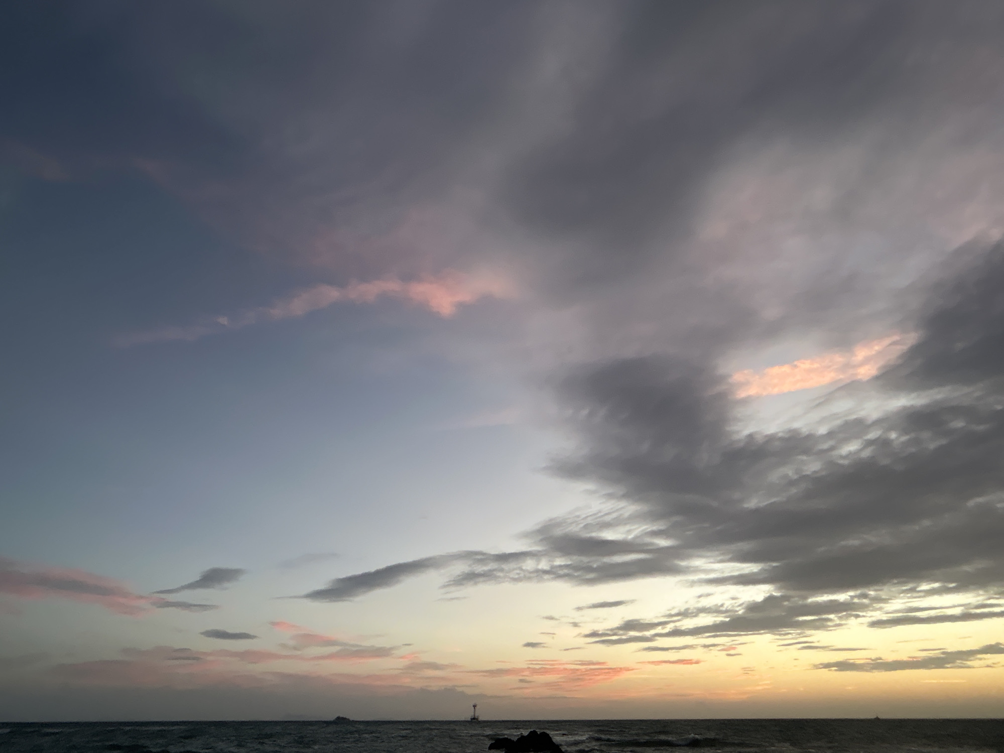 A serene seascape is captured at dusk in Ko Pha-ngan District, Thailand. The sky transitions from a soft, pale blue on the left to a warm gradient of yellow and orange on the right, where the sun is setting. Wispy clouds, tinged with pink, stretch across the horizon, interspersed with darker, more substantial grey clouds. The ocean below is a deep, dark blue, with gentle waves lapping at the shore. In the distance, a small lighthouse stands on a rocky outcrop, serving as a solitary beacon against the vast expanse of sea and sky. The overall atmosphere is tranquil and peaceful, evoking a sense of calm and natural beauty.