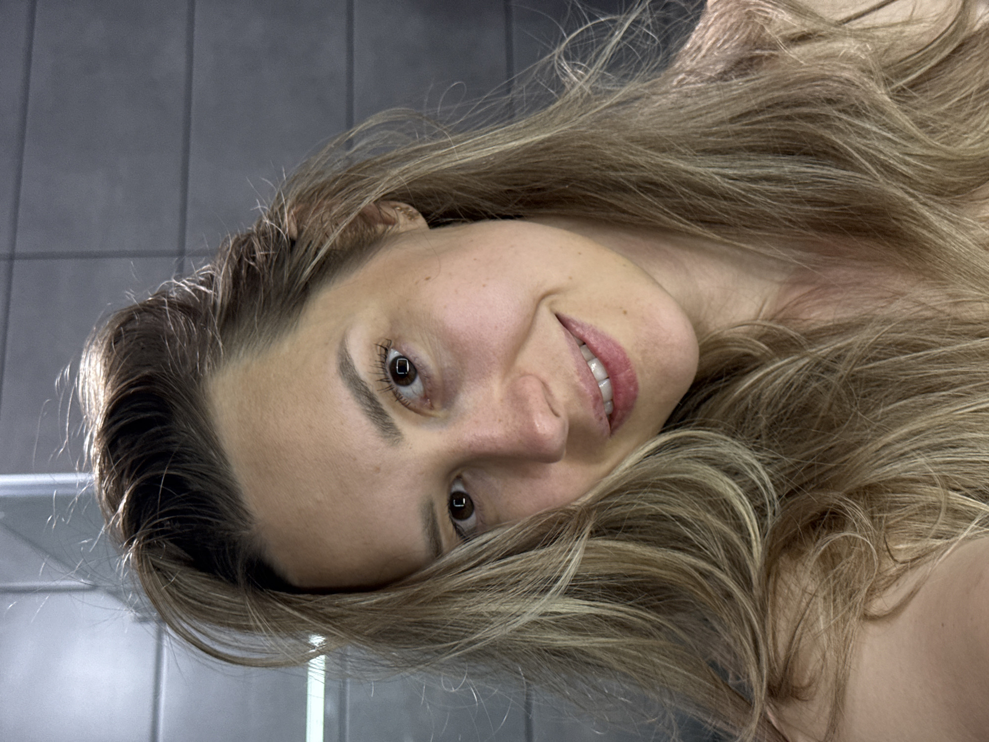 A close-up, tilted portrait captures a woman with long, wavy, blonde and light brown hair, appearing to be taken indoors. Her head is angled, with her eyes looking directly towards the viewer. She has a gentle, natural smile, revealing her teeth. Her eyes are brown, and her eyelashes show mascara. Her skin appears clear, with natural eyebrows. Her hair flows around her face and extends into the foreground and background, giving the impression of movement.
The background consists of grey, possibly tiled, walls with visible grout lines forming a grid pattern, suggesting an interior space like a modern bathroom or room. The lighting appears soft and even, typical of indoor artificial light or diffuse natural light. The overall mood is relaxed and inviting, conveyed by her soft expression. No specific location cues within the image itself confirm the provided context of Chaikovsky, Russia, beyond it being an indoor setting. There is no visible text.