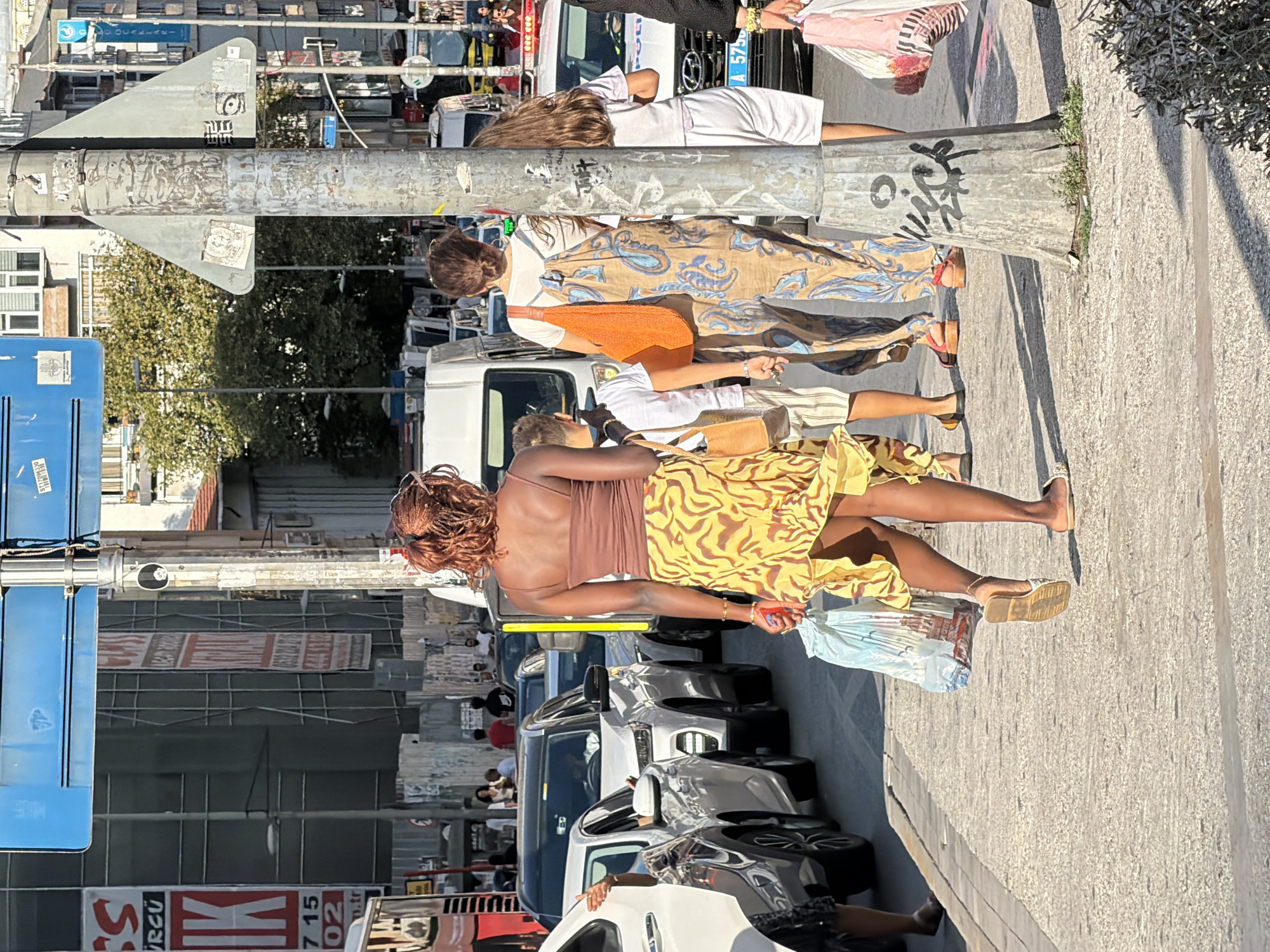 In Kadıköy, Turkey, on a sunlit city street, a group of people walks along a sidewalk bordering a line of cars. A woman in the foreground, facing away from the camera, wears a brown tank top and a yellow patterned skirt, carrying a plastic bag. Behind her, a person in a patterned jumpsuit walks with a young boy in a white t-shirt and striped shorts. Further back, a young girl in white shorts and a t-shirt walks beside a person in a white shirt. Cars are stopped in traffic, and a police car with flashing lights is visible. Buildings and trees line the street. A metal pole with graffiti stands prominently in the foreground.