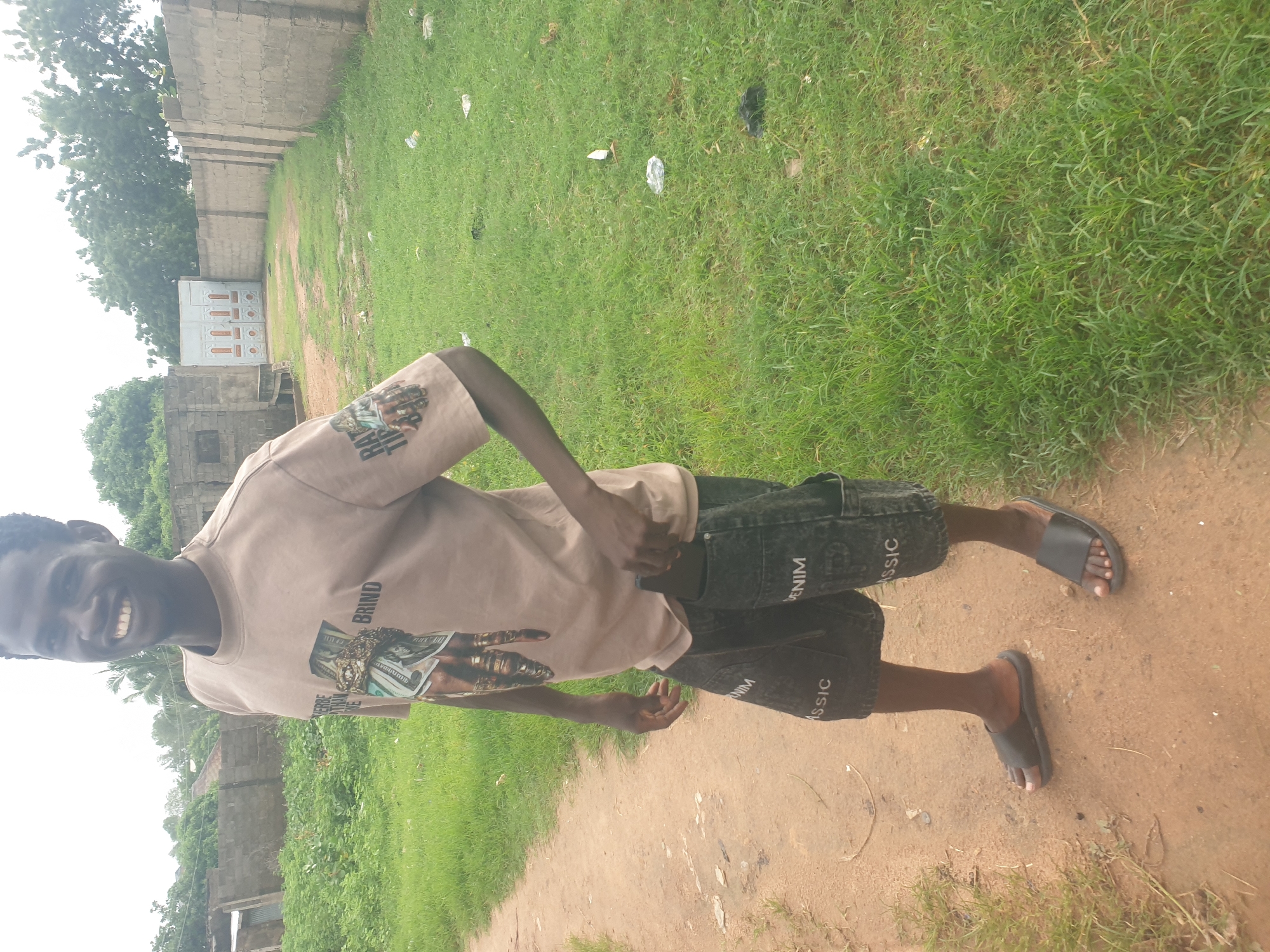 A young Black man stands on a dirt path in front of a grassy area with a wall and buildings in the background. He is wearing a light brown t-shirt with a graphic of money on it, dark denim shorts with "DENIM" and "CLASSIC" printed on them, and black slide sandals. His right hand is in his shorts pocket, and he is smiling at the camera. The environment appears to be an outdoor area in a residential or industrial part of a city, with some litter visible in the grass. The sky is overcast.