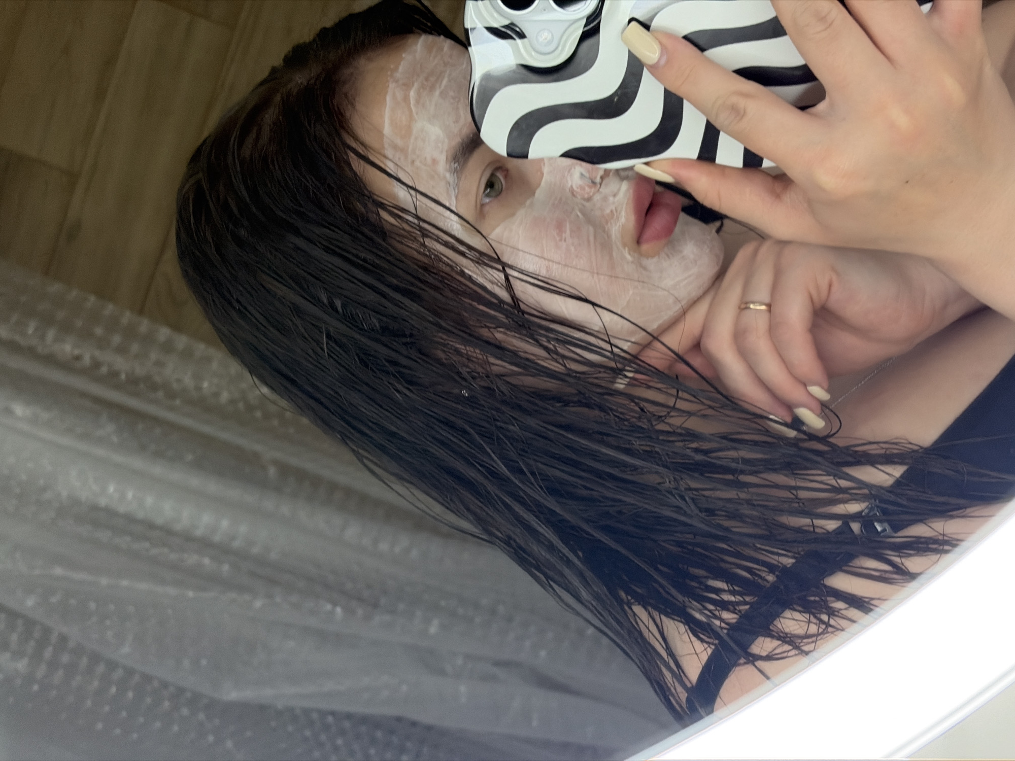 A woman is taking a selfie indoors, possibly in Krasnodar, Russia, given the information. She is wearing a face mask and holding a smartphone with a black and white patterned case. Her dark hair is wet. She is wearing a black top. A ring is visible on one of her fingers. The background includes a reflective surface, possibly a mirror, and a white, textured material.
